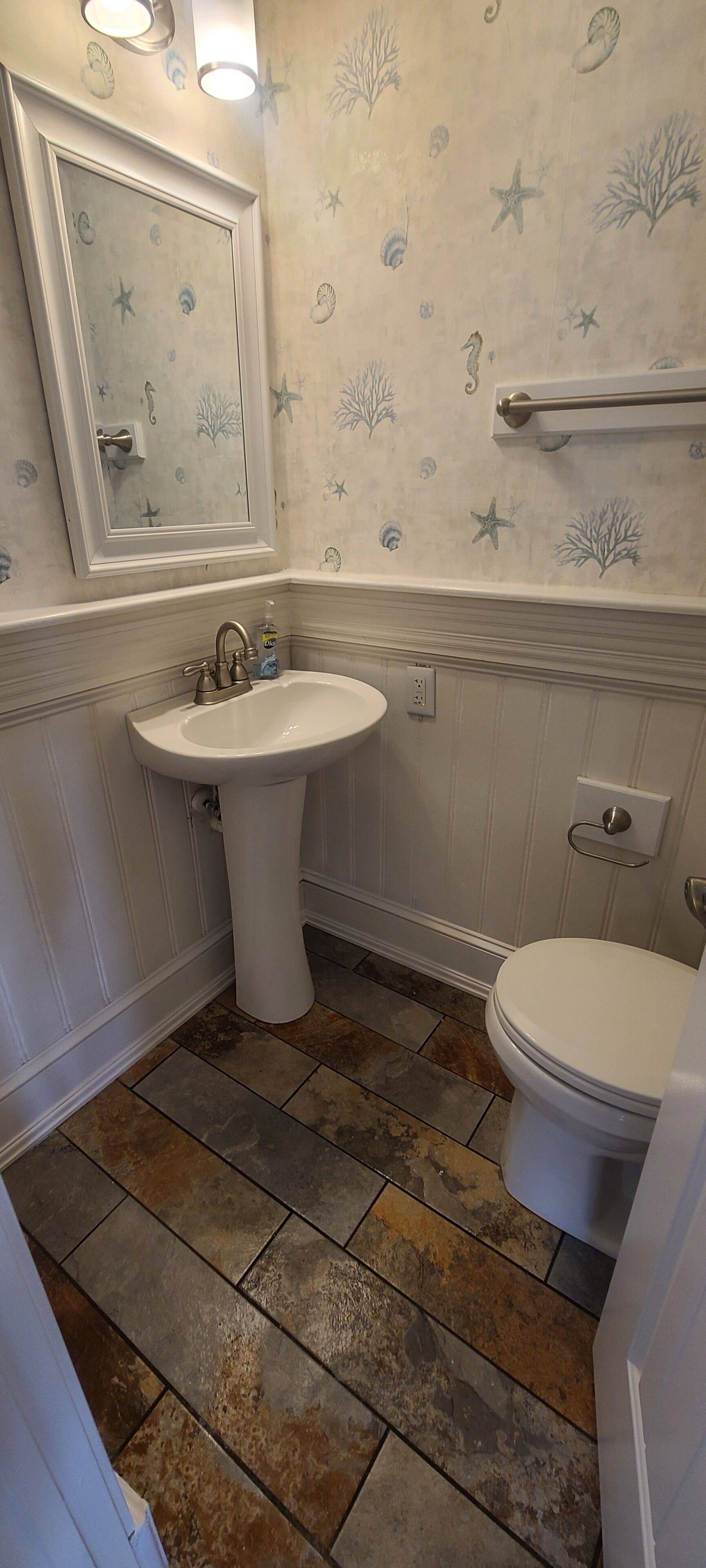 Une salle de bain avec un lavabo blanc, des toilettes et un miroir au-dessus du lavabo. Les murs sont recouverts d’un papier peint sur le thème de la mer.
