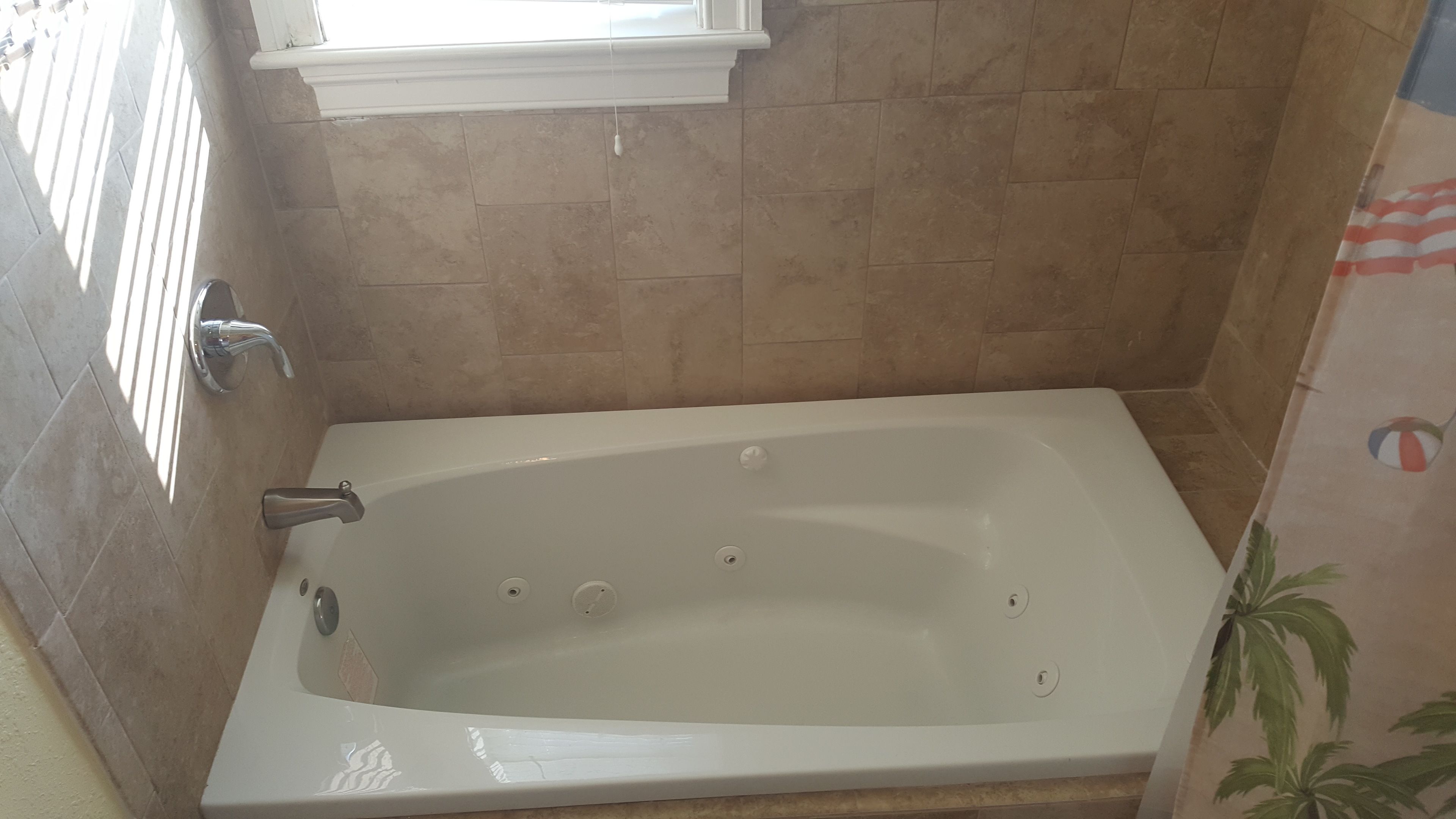 Un baño con una bañera blanca, paredes de baldosas color beige y una ventana sobre la bañera.