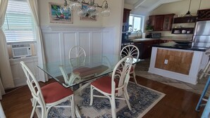 A dining area with a glass table and white chairs, a kitchen in the background, and a patterned rug on the floor. - Pierhouse Bed & Breakfast (Kill Devil Hills)
