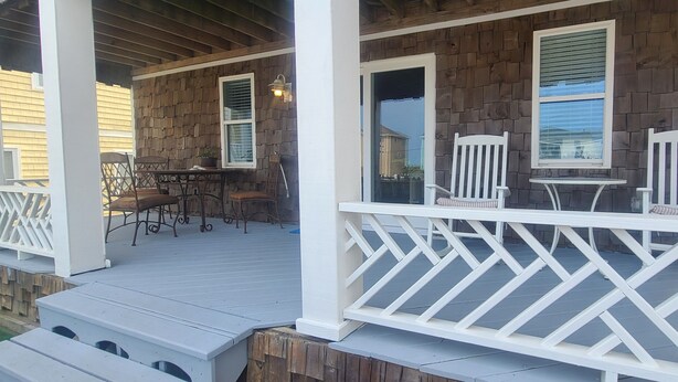 A covered porch with a wooden deck, white railings, and outdoor furniture including chairs and a table. - Pierhouse Bed & Breakfast (Kill Devil Hills)