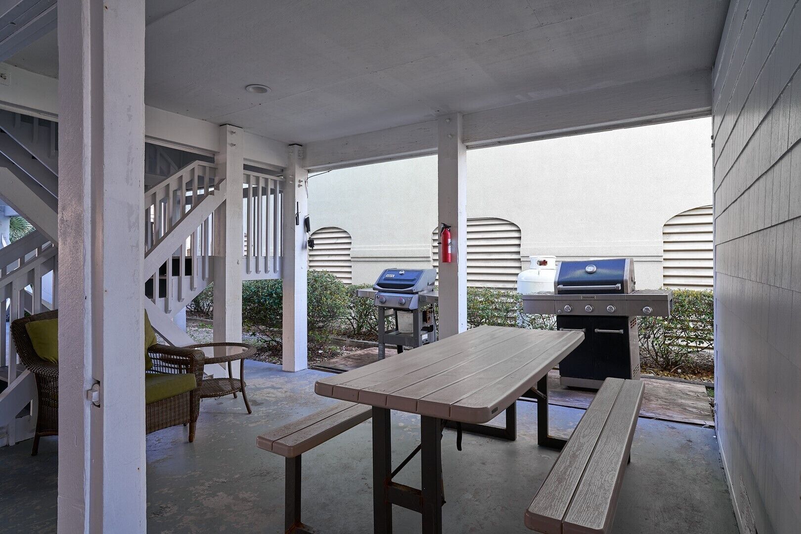 An outdoor patio with a wooden table and benches, a grill, and wicker chairs.