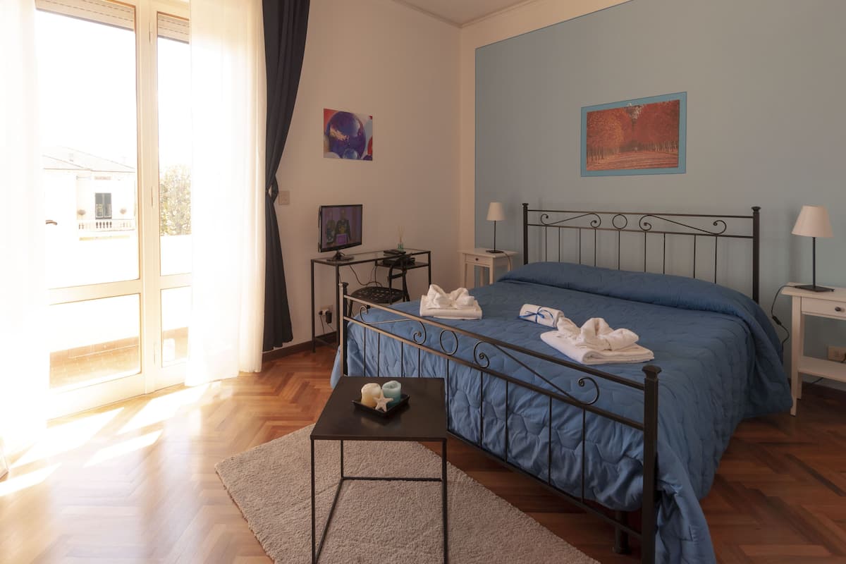 Un dormitorio con colcha azul, una mesita con velas, un escritorio con computadora y una ventana con cortinas.