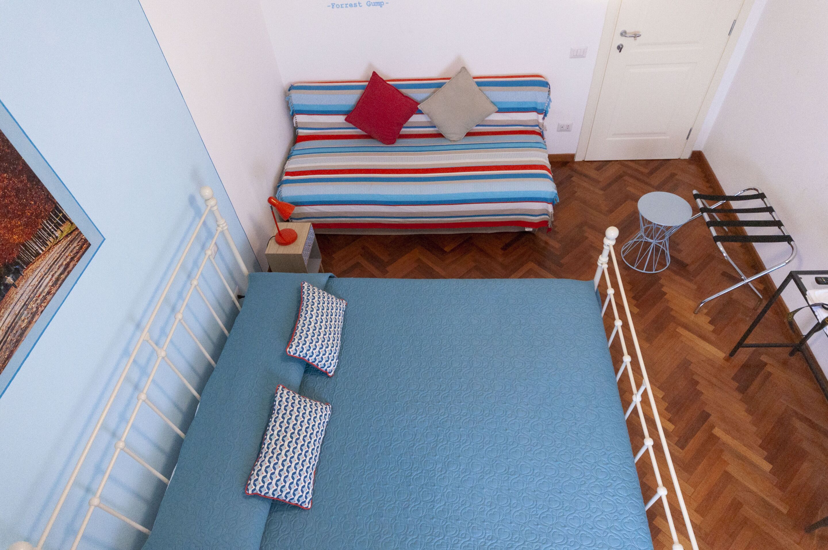 A small, cozy room with a striped bed, two patterned pillows, a small table with a bottle, and a chair with a metal frame.