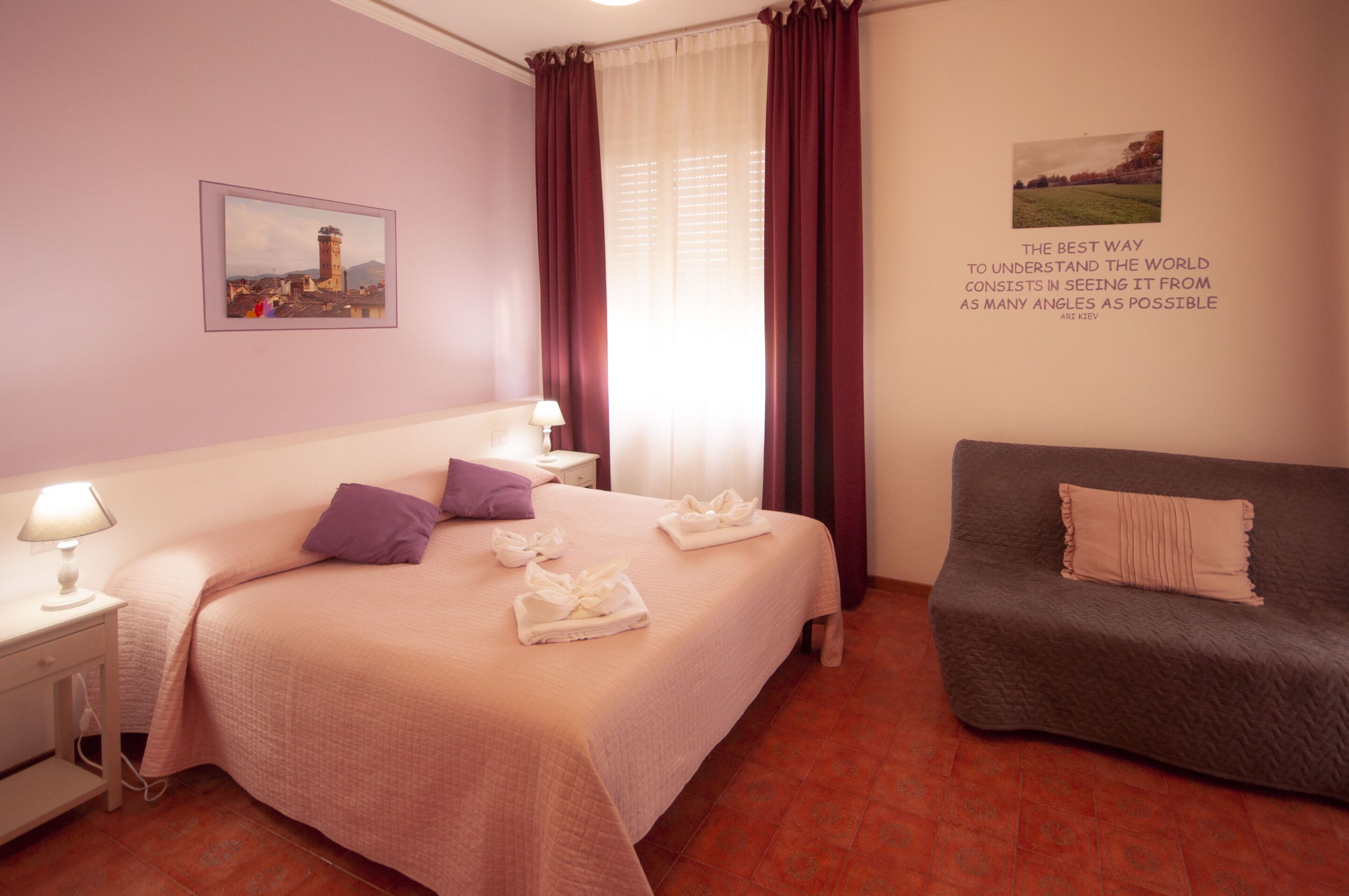 A hotel room with a bed, a nightstand, a lamp, a sofa, and two framed pictures on the walls.