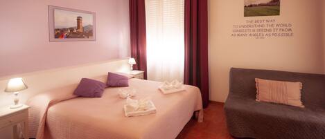 A hotel room with a bed, a nightstand, a lamp, a sofa, and two framed pictures on the walls.