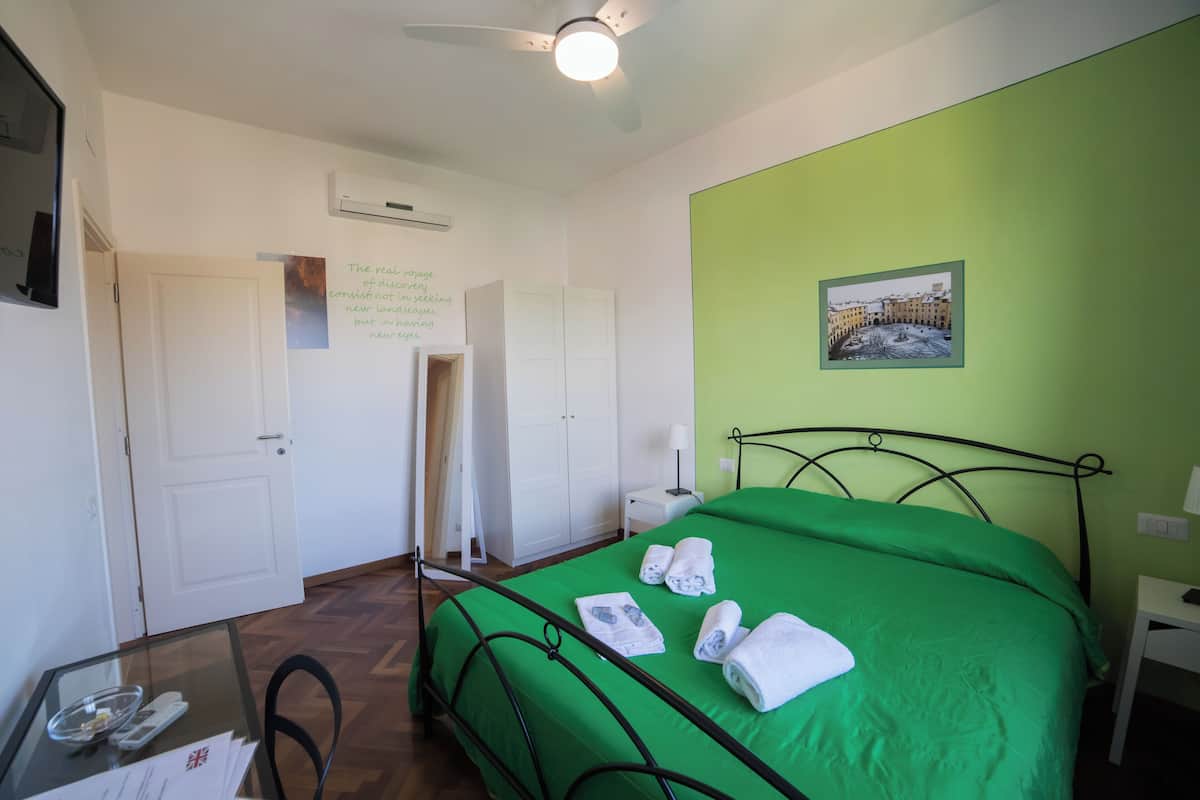 Una habitación con una cama verde, paredes blancas, un ventilador de techo, un cuadro enmarcado y una puerta.