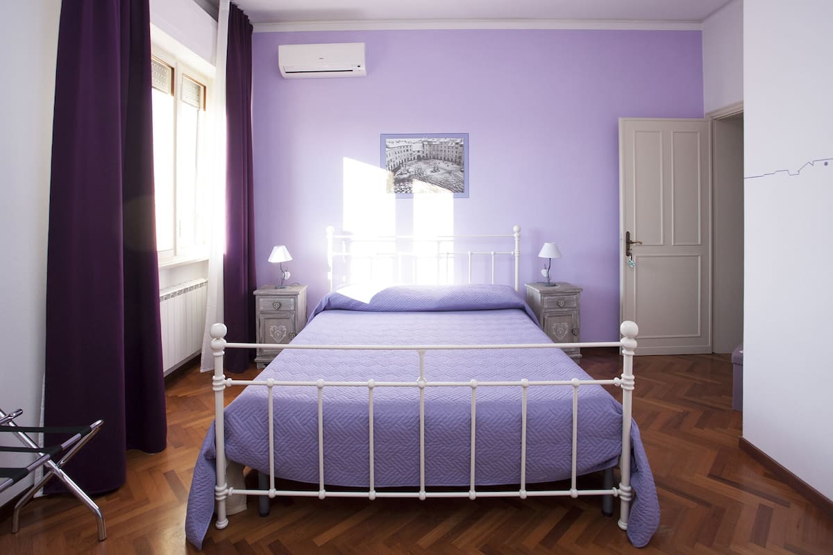 Un dormitorio con una estructura de cama de metal blanco, ropa de cama morada, dos mesitas de noche con lámparas, una ventana con cortinas moradas y una puerta.