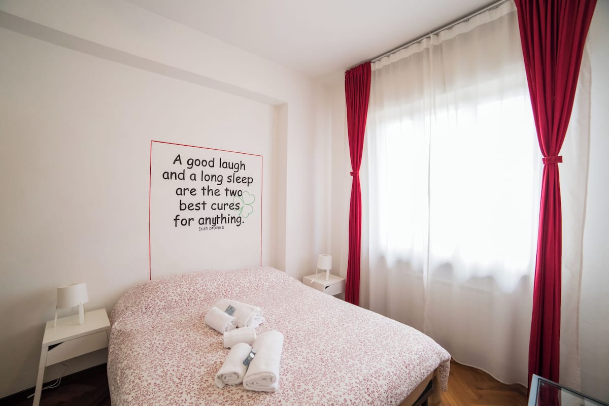 Un dormitorio con cama, mesita de noche con lámpara, pared con cita y cortinas rojas.