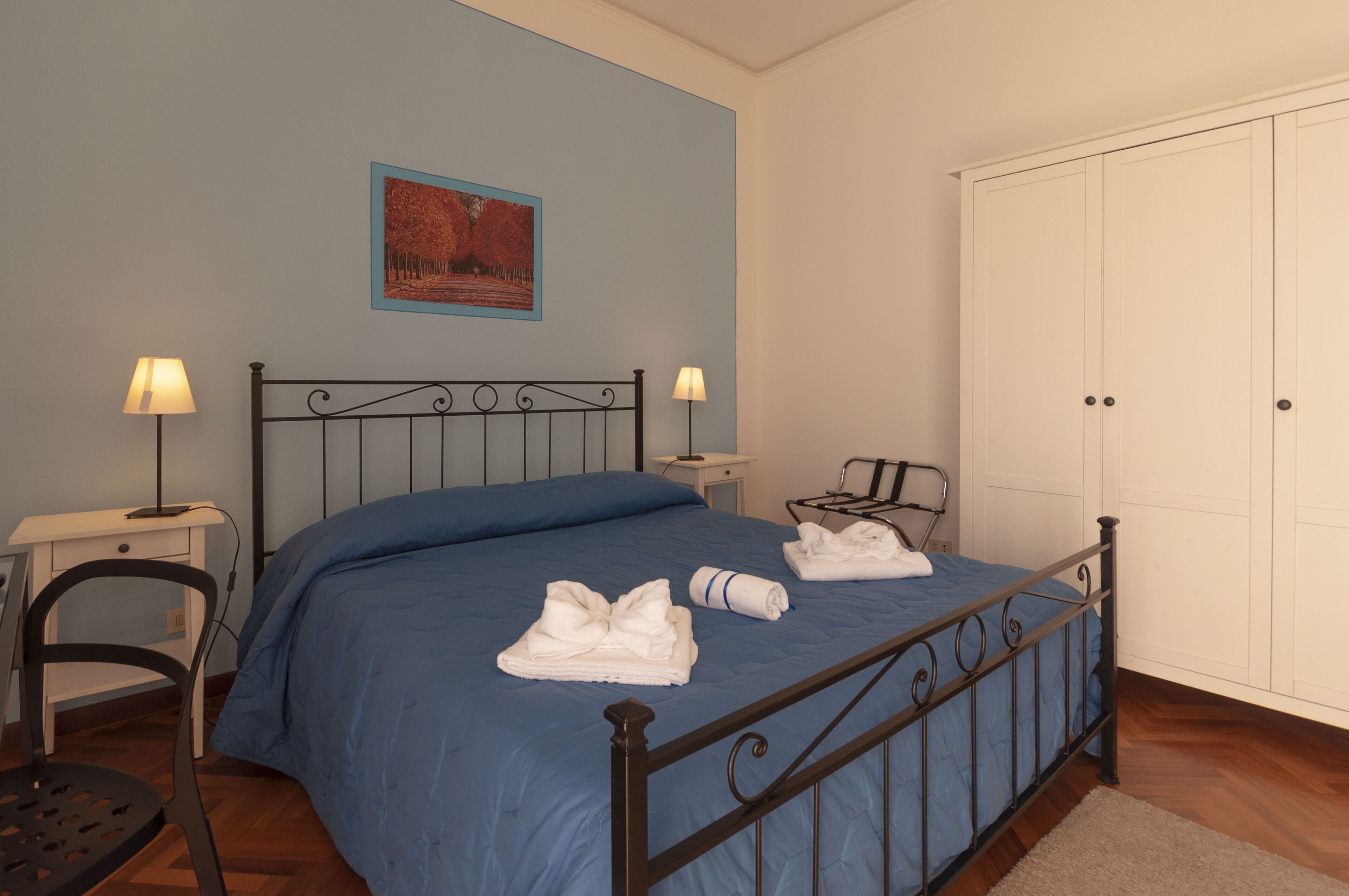 A neatly made bed with blue bedding, two bedside lamps, a chair, a desk, and a wardrobe.