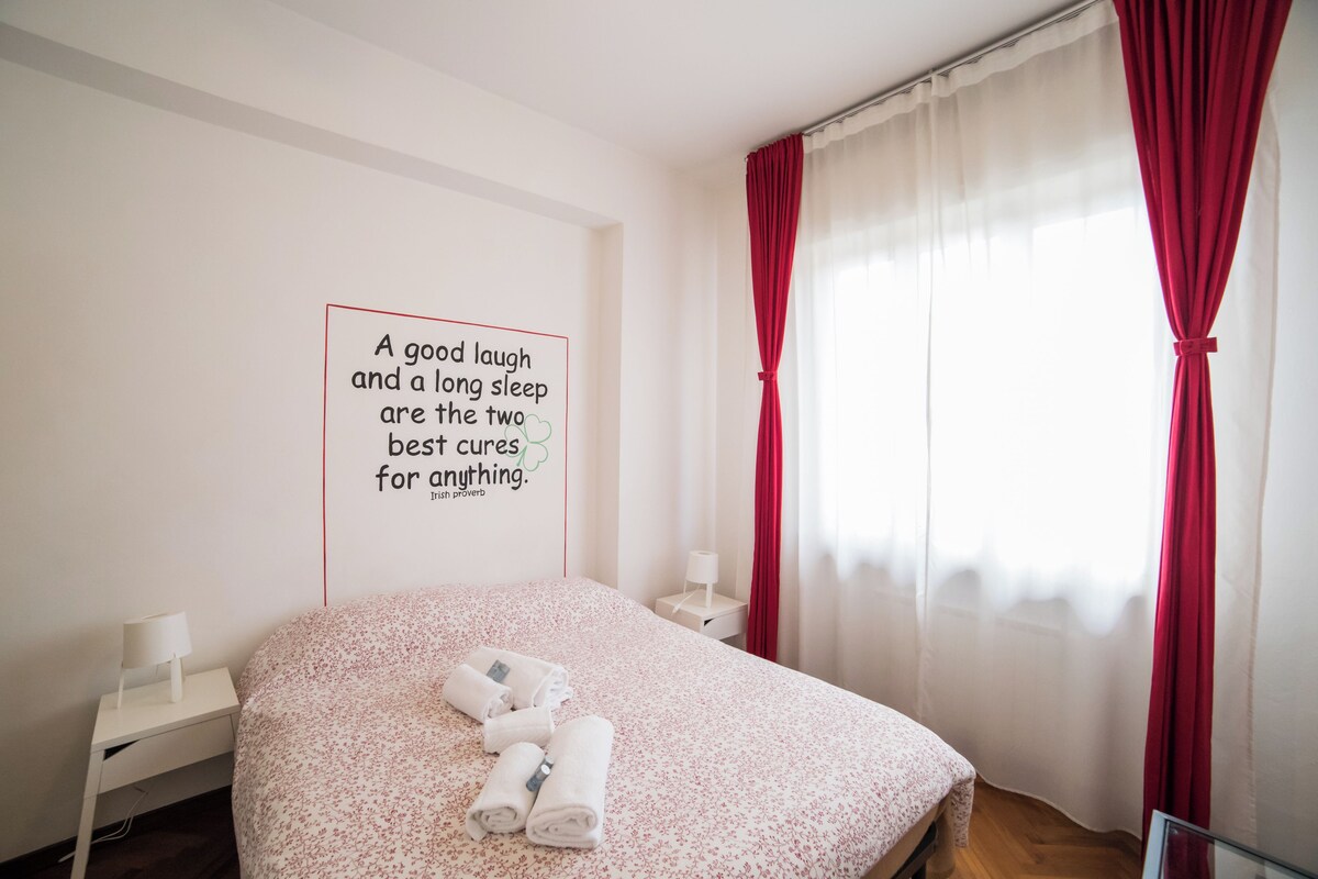 Un dormitorio con cama, mesita de noche con lámpara, pared con cita y cortinas rojas.