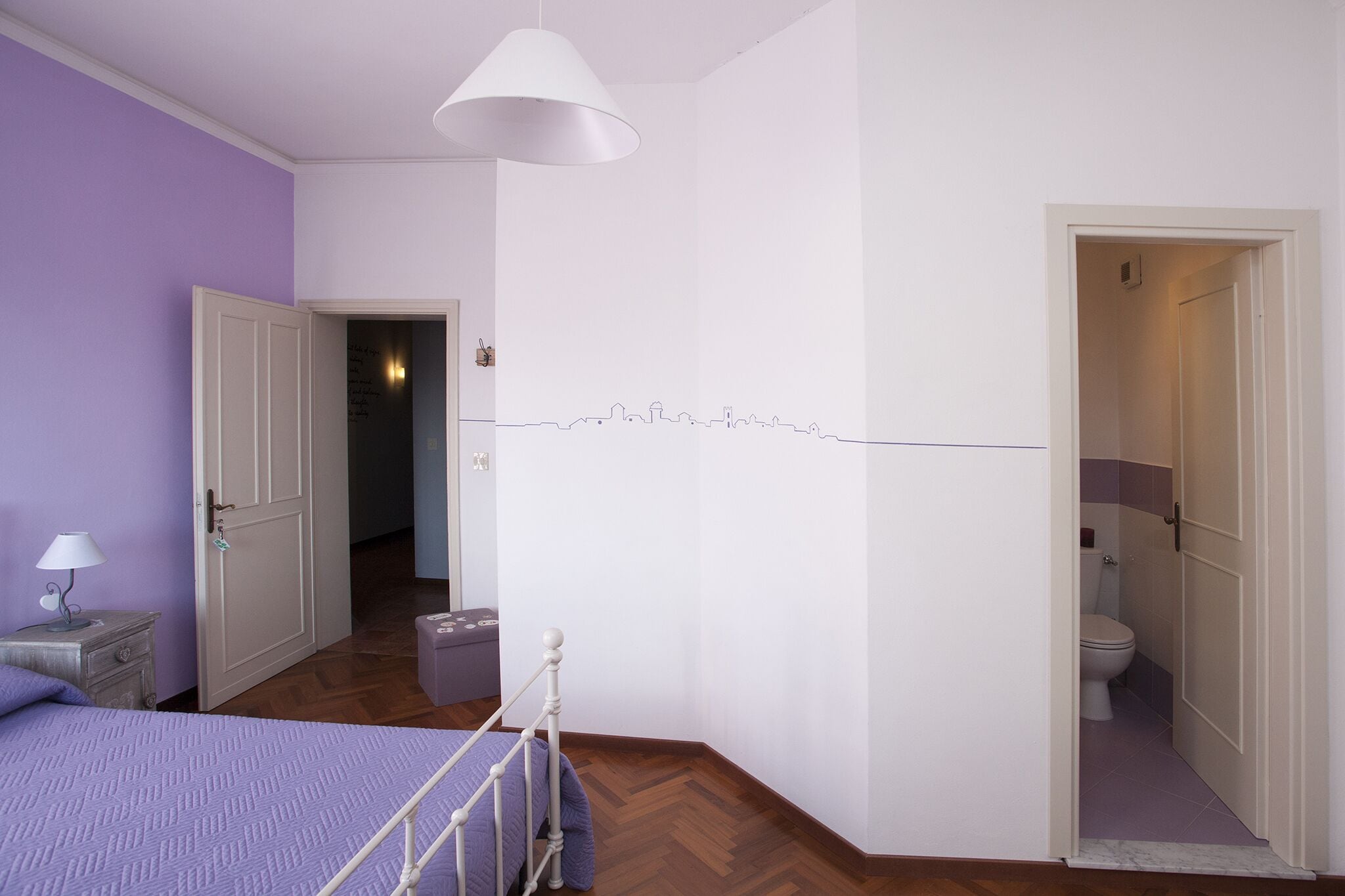 A bedroom with a bed, a nightstand, a lamp, and a bathroom visible through an open door.