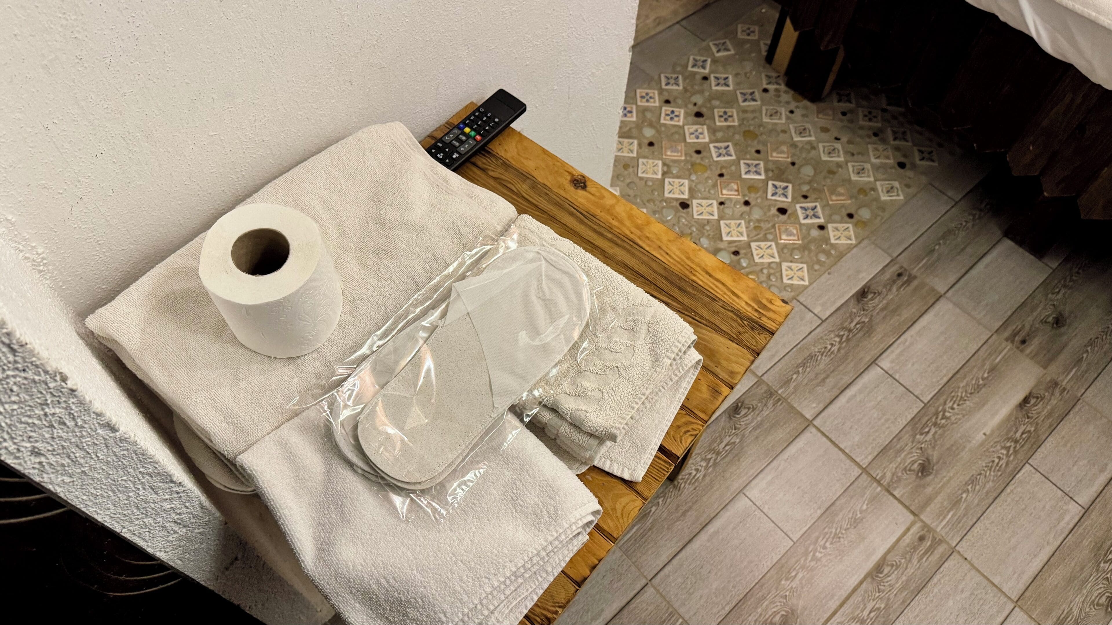 Un baño con suelo de madera, una toalla, un rollo de papel higiénico y un mando a distancia sobre una superficie de madera.