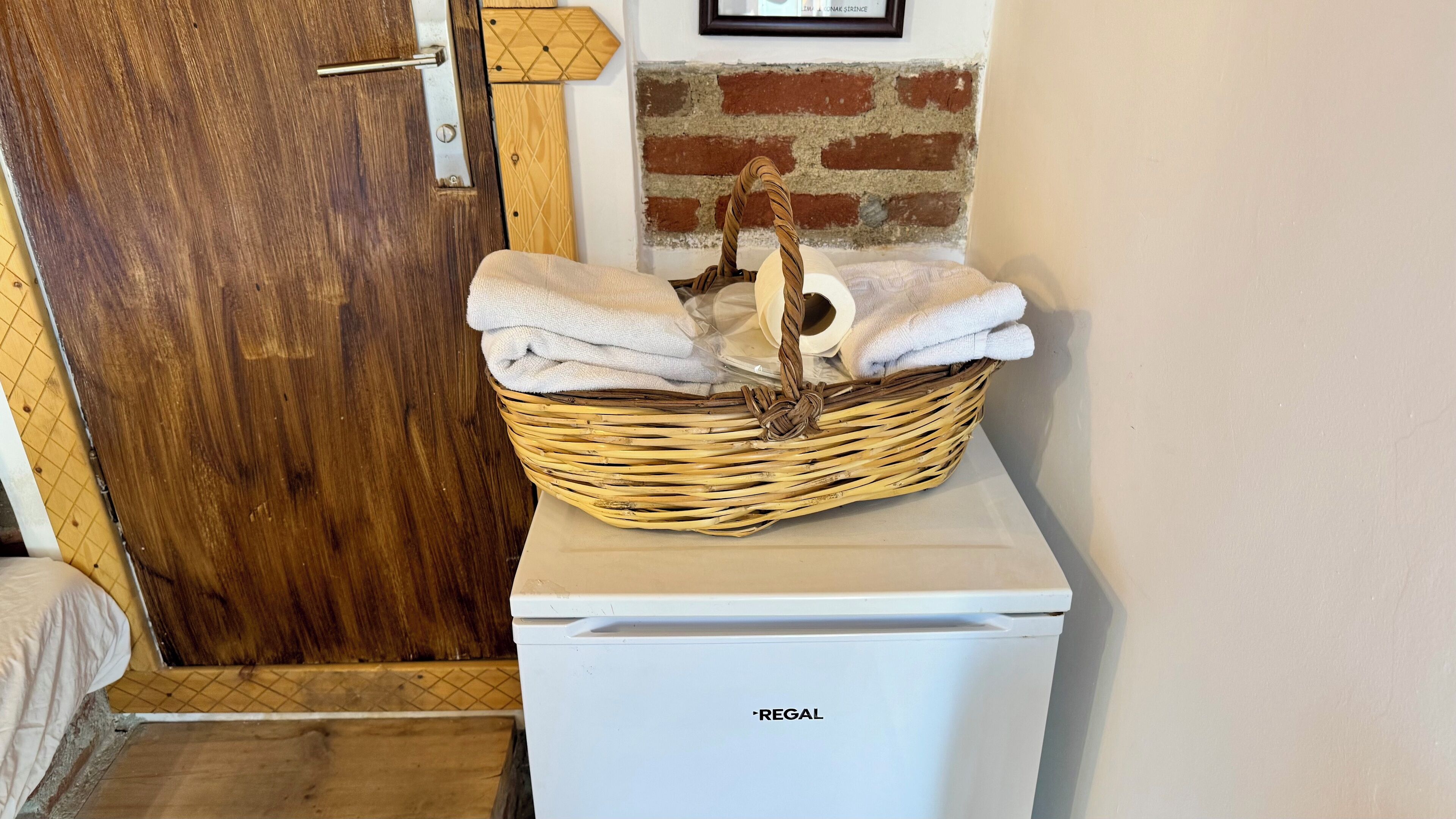 A white refrigerator with the brand name 'REGAL' on it, placed in a room with a wooden door and a brick wall.