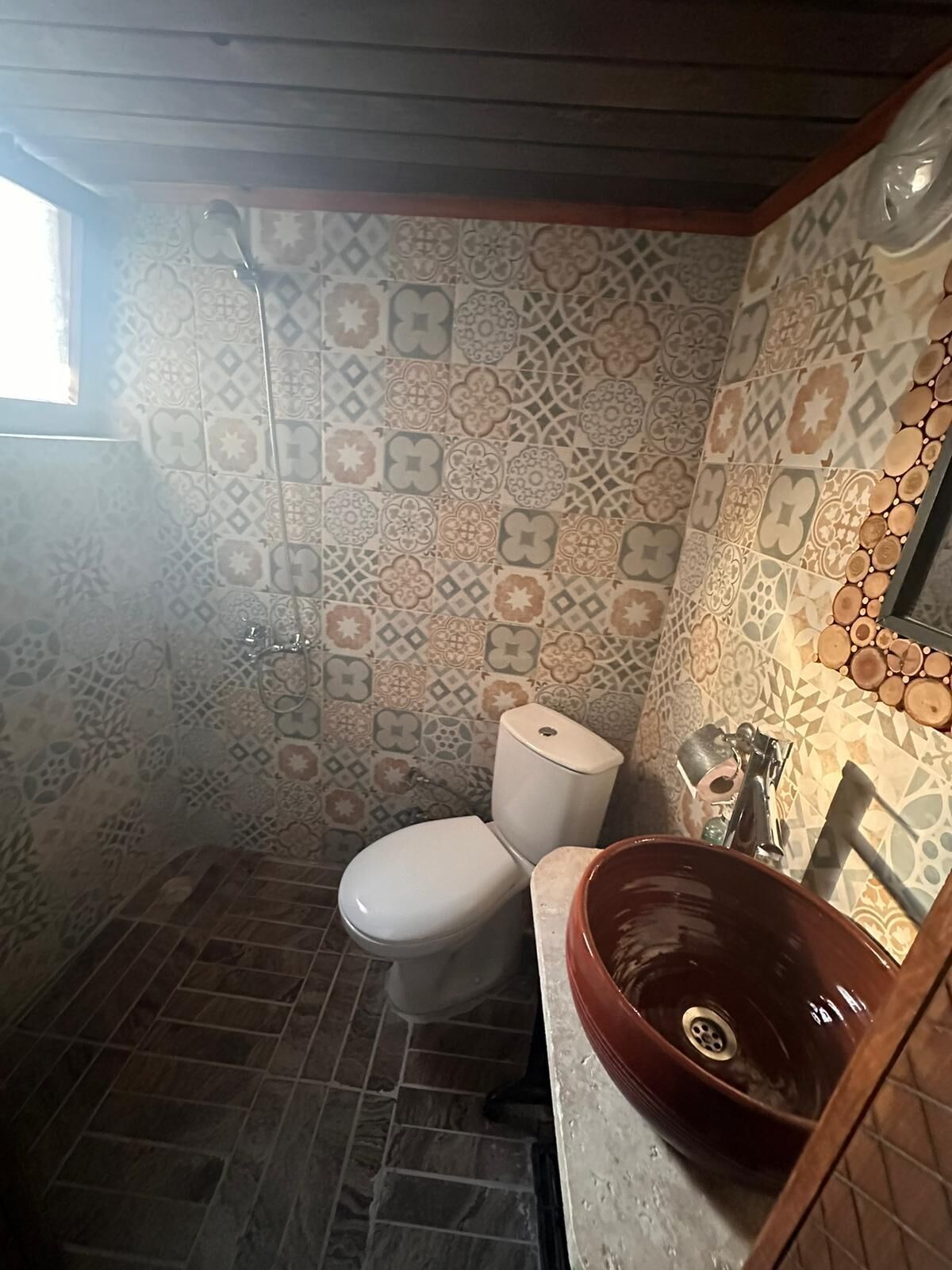 Un baño con baldosas estampadas, inodoro blanco, lavamanos marrón y ducha.