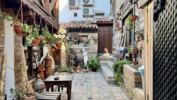 A narrow alleyway with a rustic stone floor, potted plants, a wooden table, and a statue. - Limaki Konak Sirince (Selçuk)
