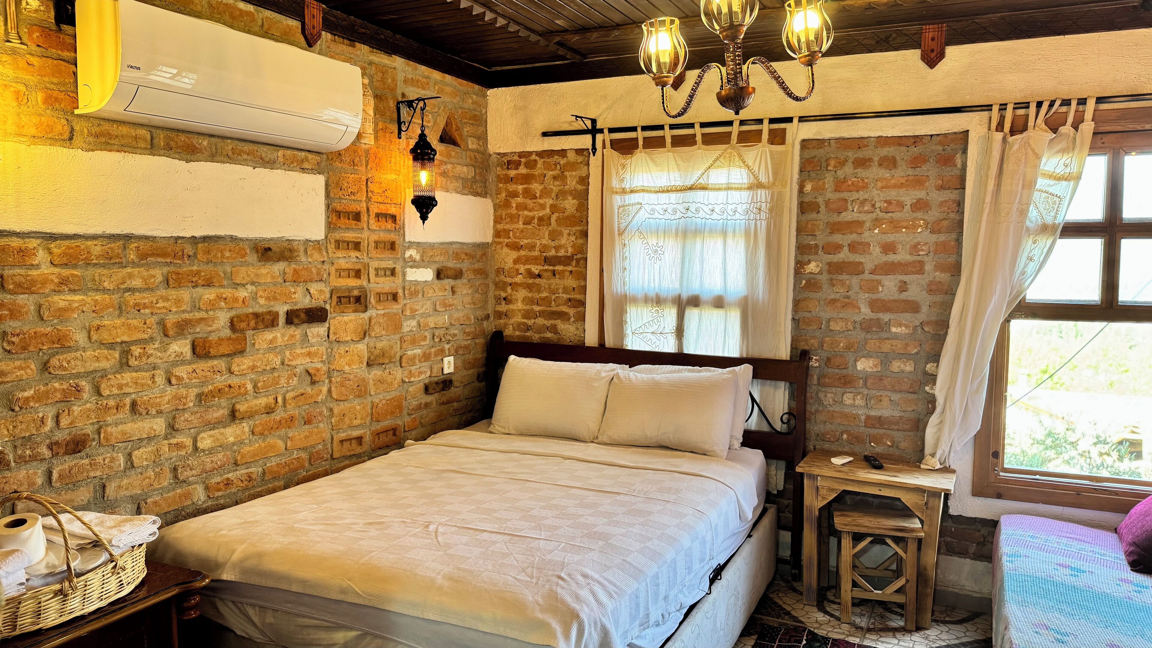 A cozy room with a brick wall, a bed with white bedding, a small wooden nightstand, a wicker basket, and a hanging lantern.