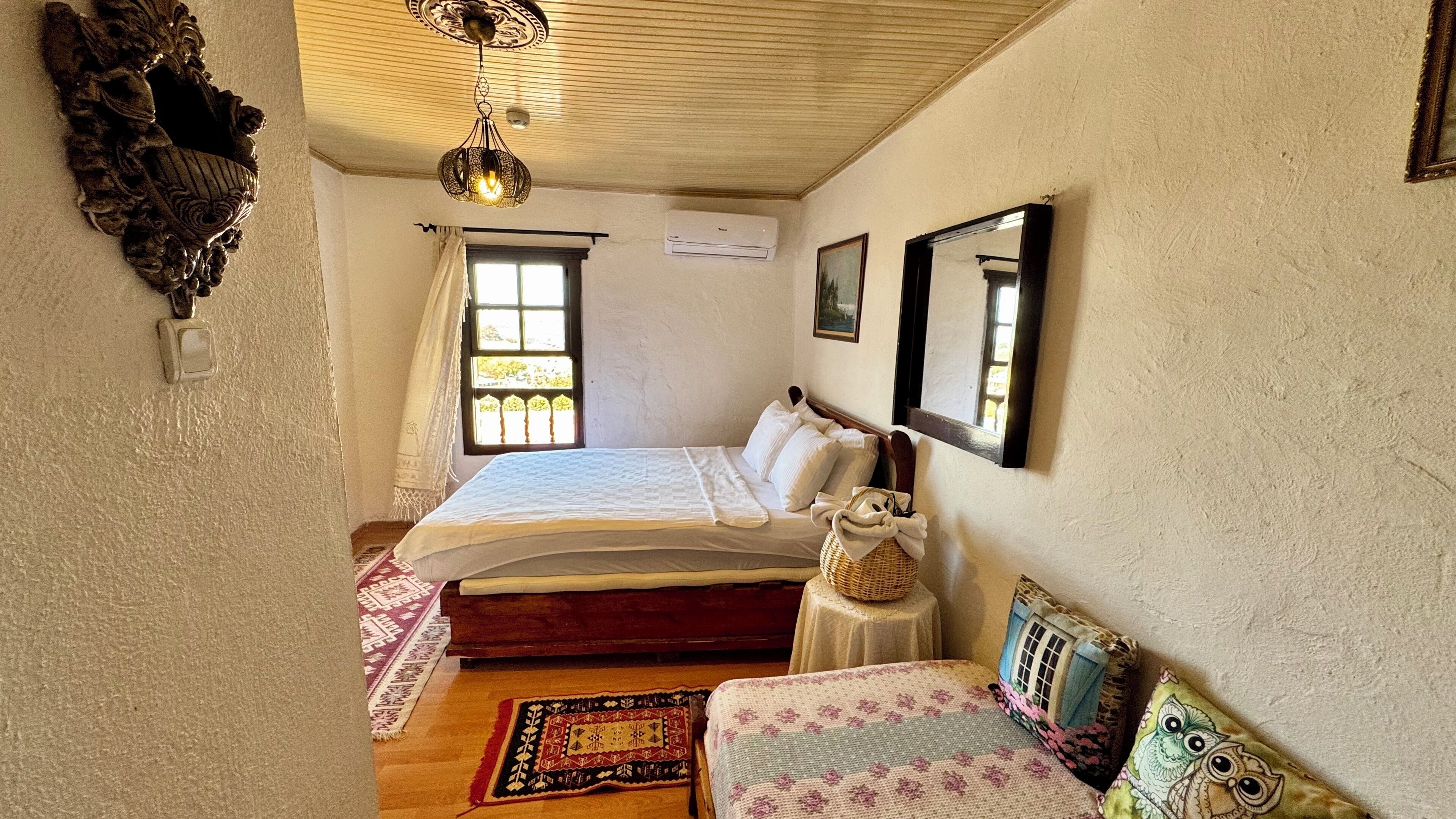 A bedroom with a bed, a bedside table with a basket, a mirror, and a window with curtains.