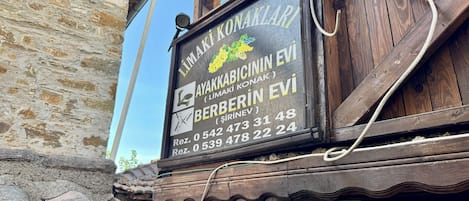 Puinen kyltti, jossa on yrityksen yhteystiedot. Yrityksen nimi on »AYAKKABICININ EVI» ja se sijaitsee alueella nimeltä »SİRİNEV».