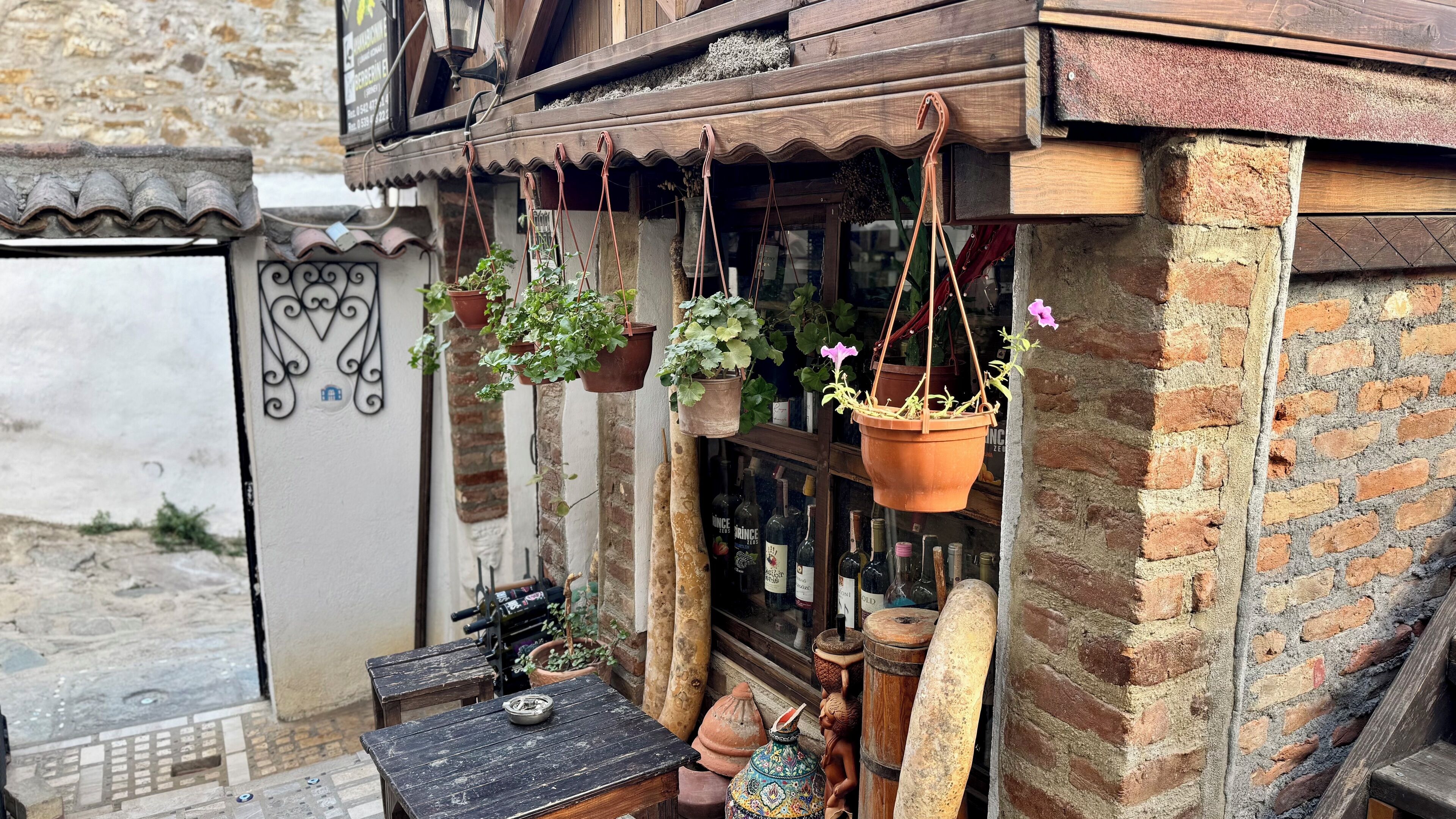 A rustic building with hanging flower pots and a wooden table with a bottle.