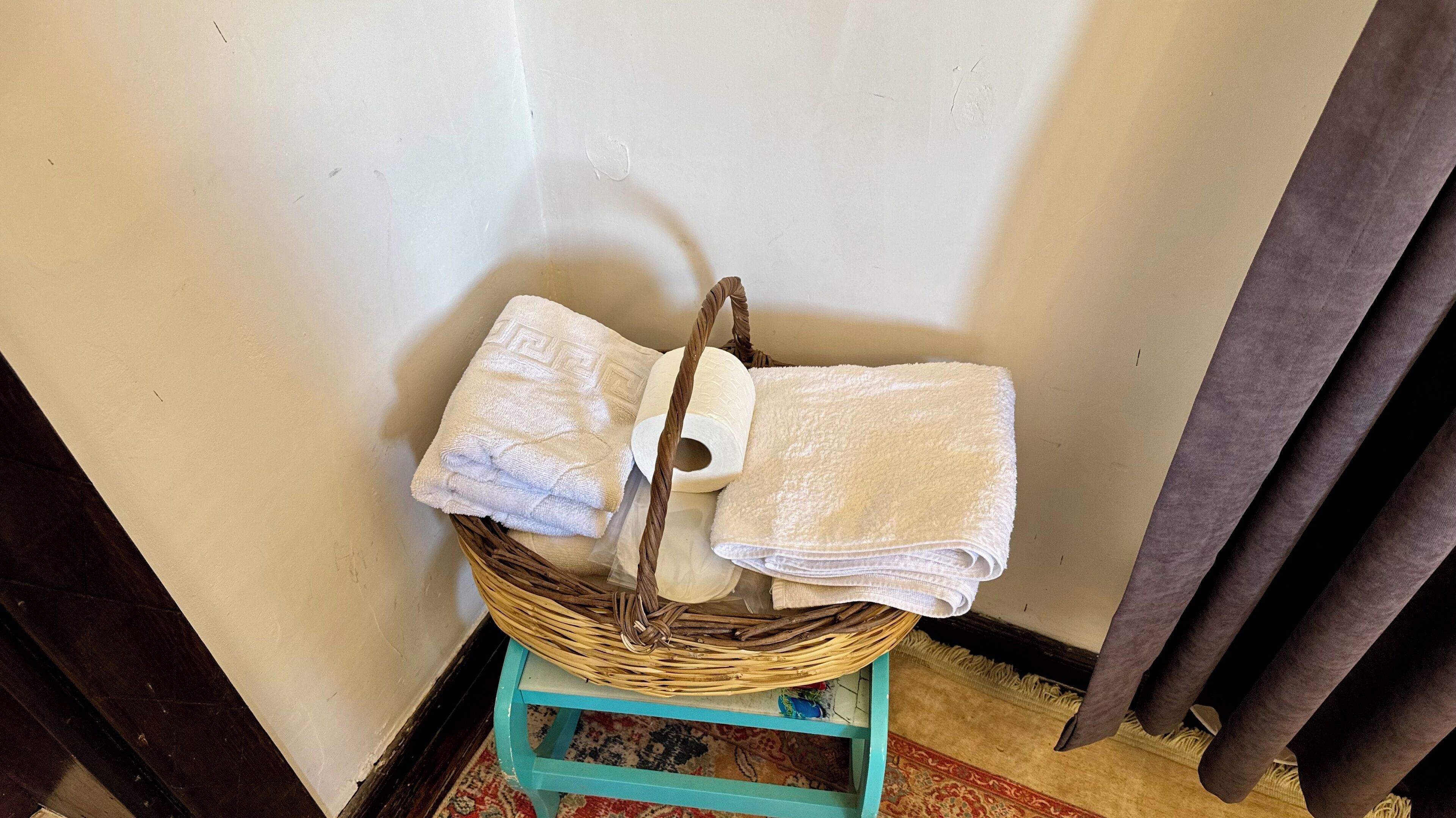 A wicker basket with towels and a roll of toilet paper on a small stool.