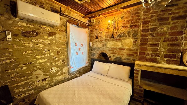 Una habitación rústica con paredes de ladrillo visto, techo de madera, cama con ropa de cama blanca, lámpara colgante y un equipo de aire acondicionado.
