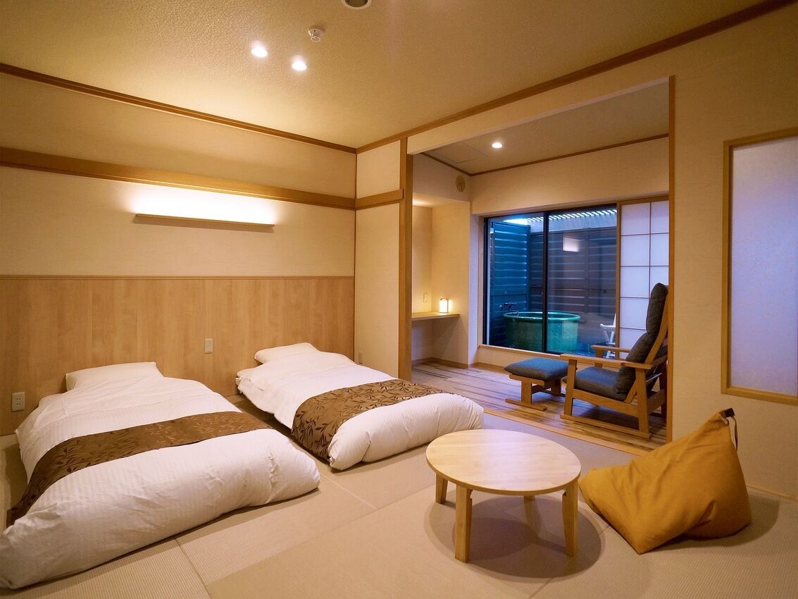 A hotel room with two beds, a small round table, a chair, and a view of the outside.
