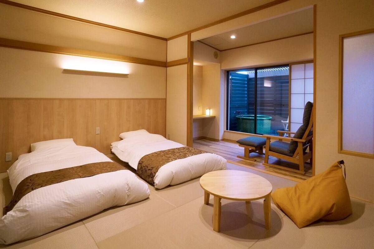 A hotel room with two beds, a small round table, a chair, and a view of the outside.