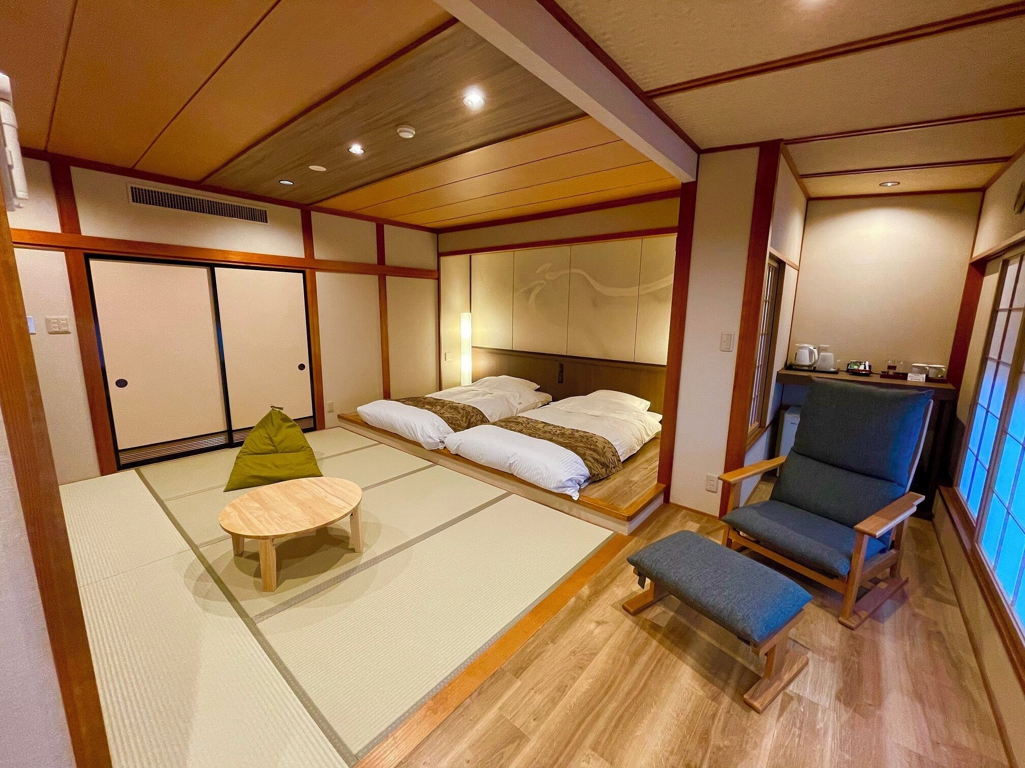 A traditional Japanese-style room with tatami flooring, a low wooden bed with white bedding, a green cushion, a wooden table, and a blue chair.
