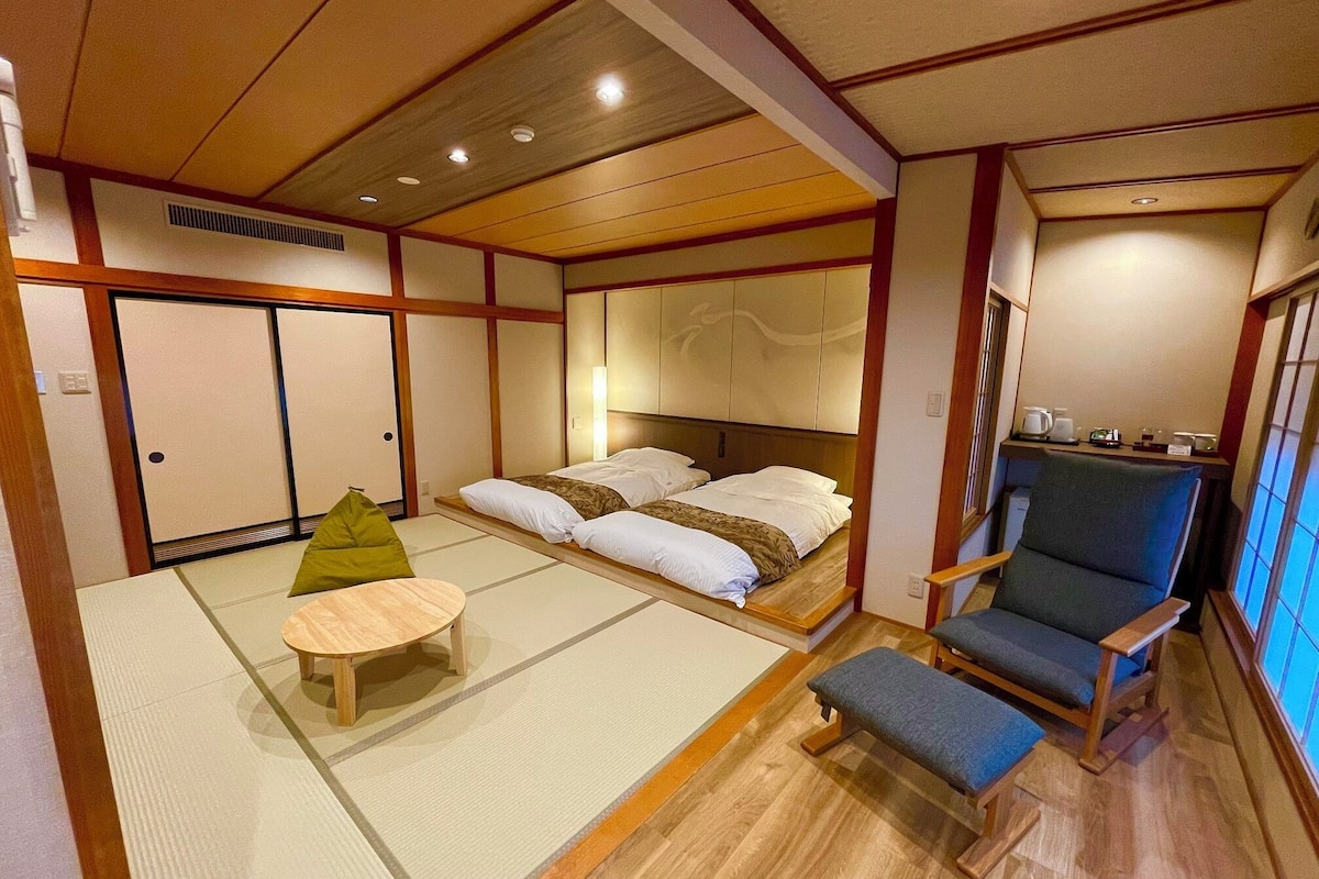 A traditional Japanese-style room with tatami flooring, a low wooden bed with white bedding, a green cushion, a wooden table, and a blue chair.