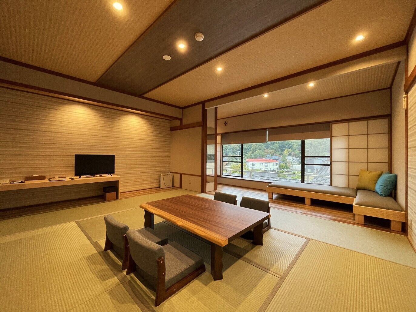 A traditional Japanese-style room with tatami flooring, a low wooden table, tatami seating, a television, and a view of a red-roofed building through large windows.