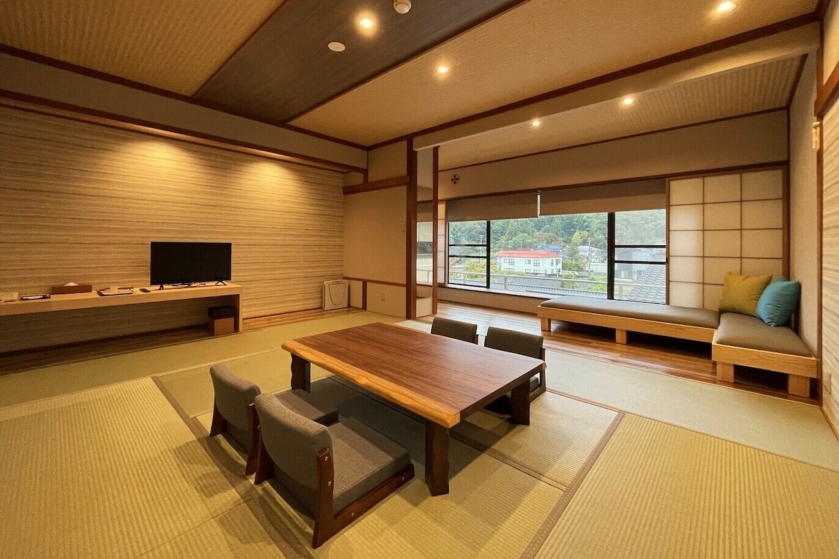 A traditional Japanese-style room with tatami flooring, a low wooden table, tatami seating, a television, and a view of a red-roofed building through large windows.