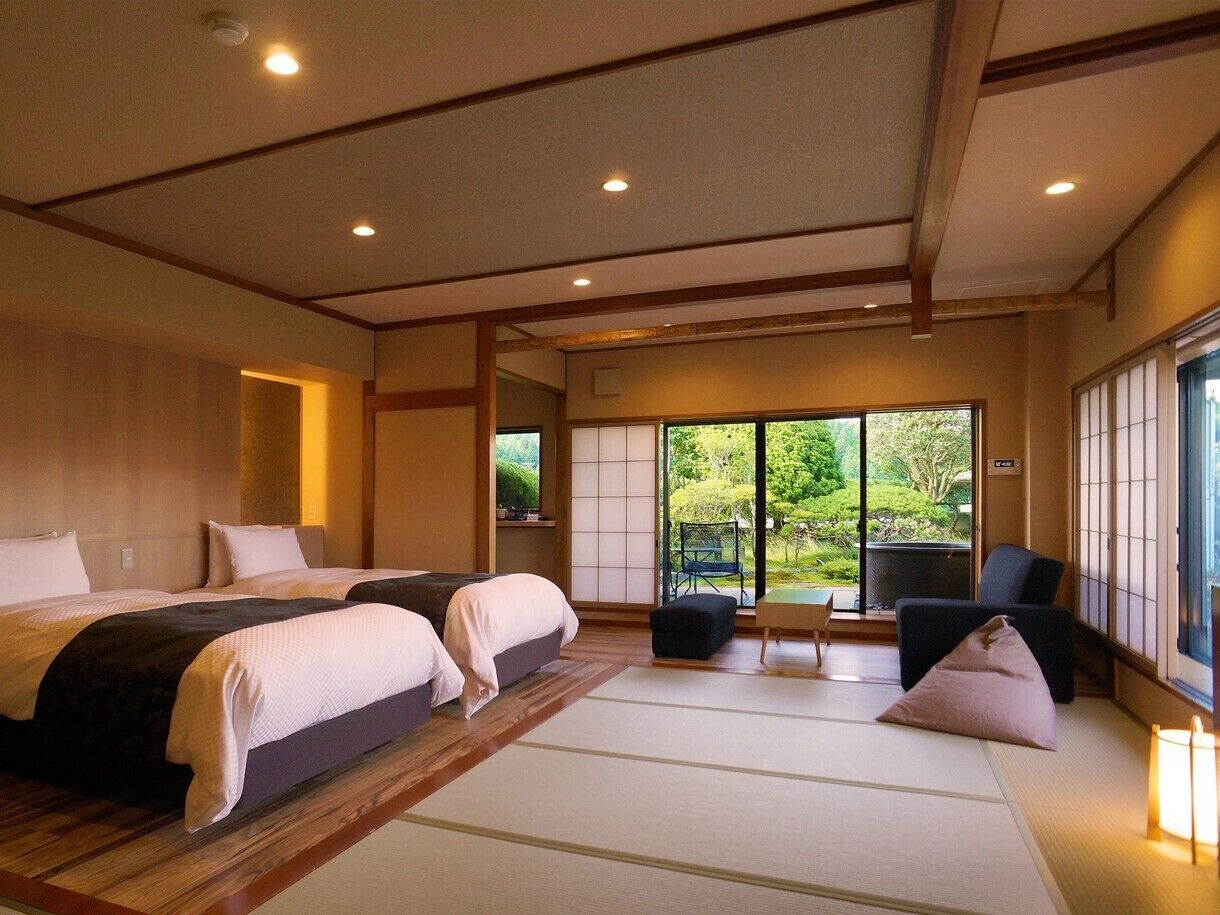 A traditional Japanese-style bedroom with tatami flooring, two beds, a sliding door to a garden, and a view of greenery.