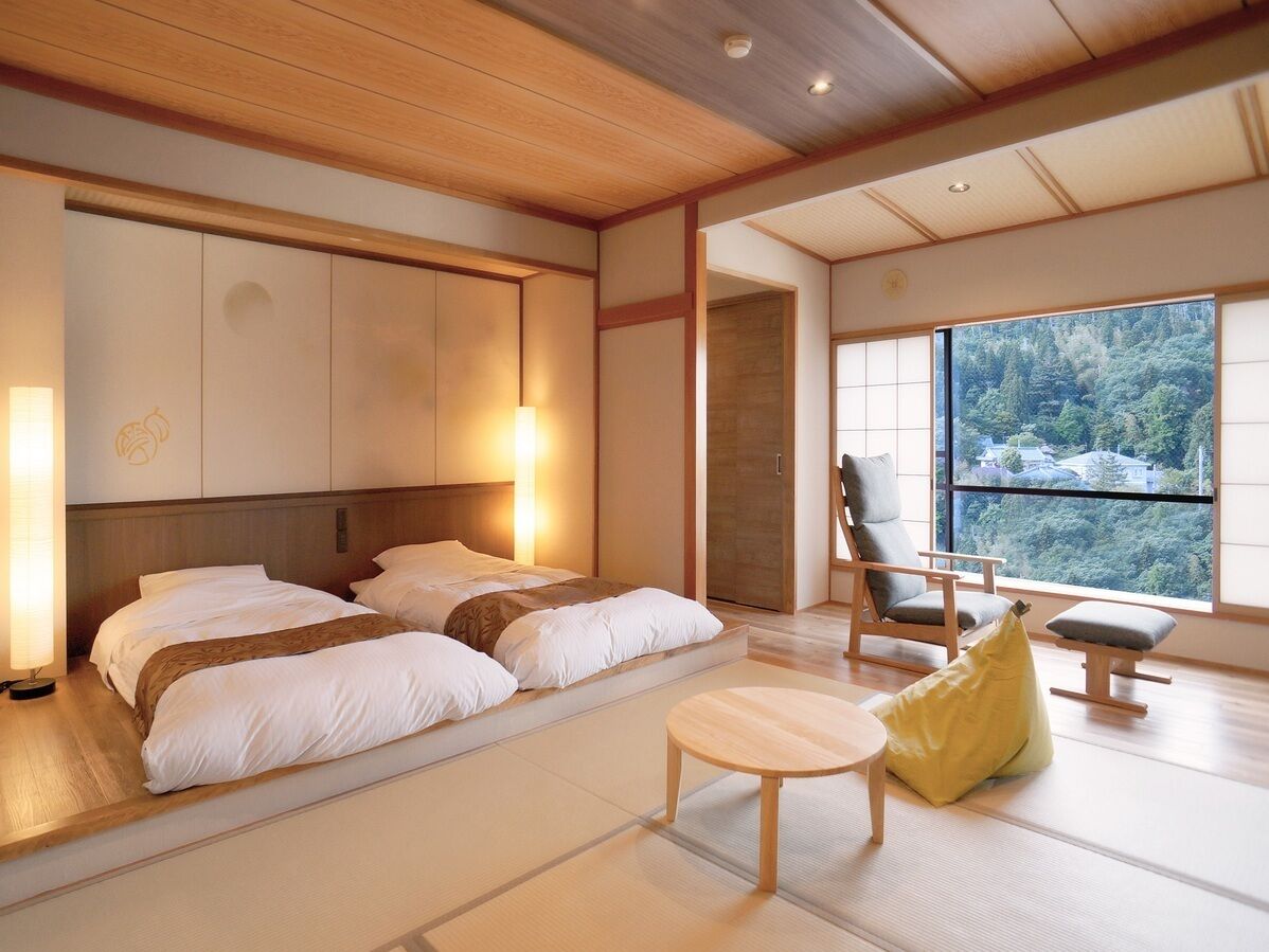 A hotel room with a bed, a chair, a small table, and a view of a forest through a large window.