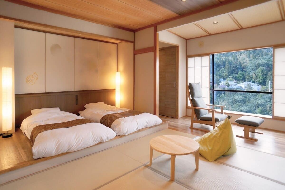 A hotel room with a bed, a chair, a small table, and a view of a forest through a large window.