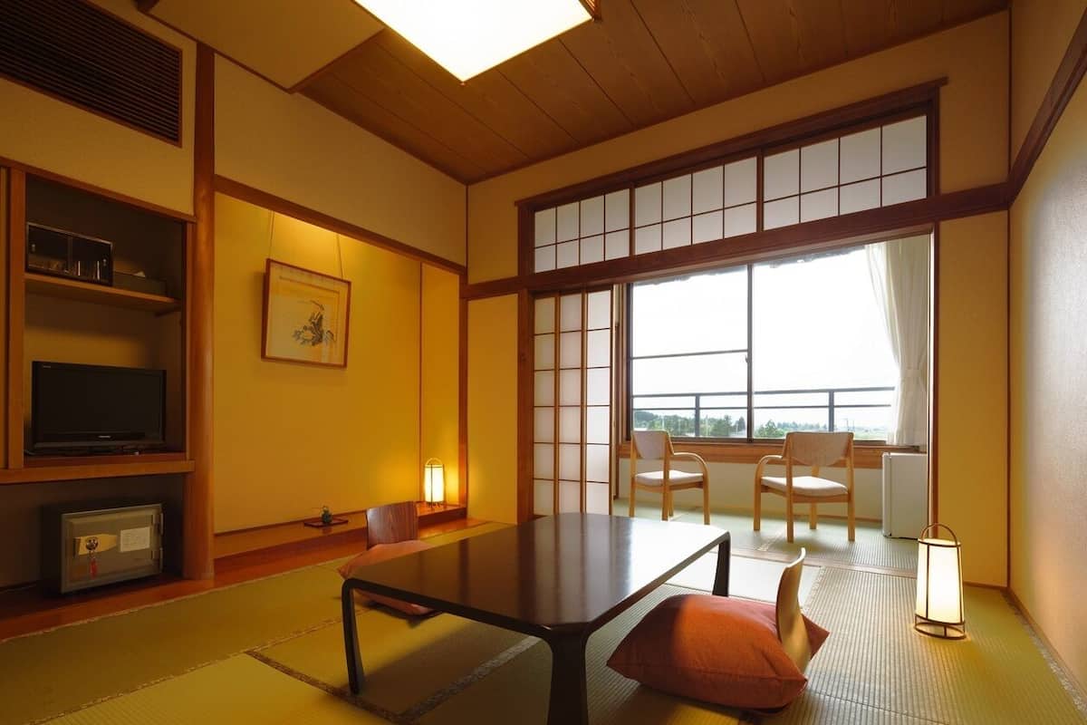 A traditional Japanese room with tatami flooring, a low table, chairs, a television, and a window with a view.