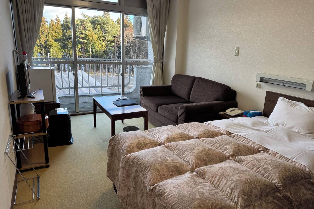 A hotel room with a bed, a sofa, a desk with a computer, a window with a view of trees, and an air conditioning unit.