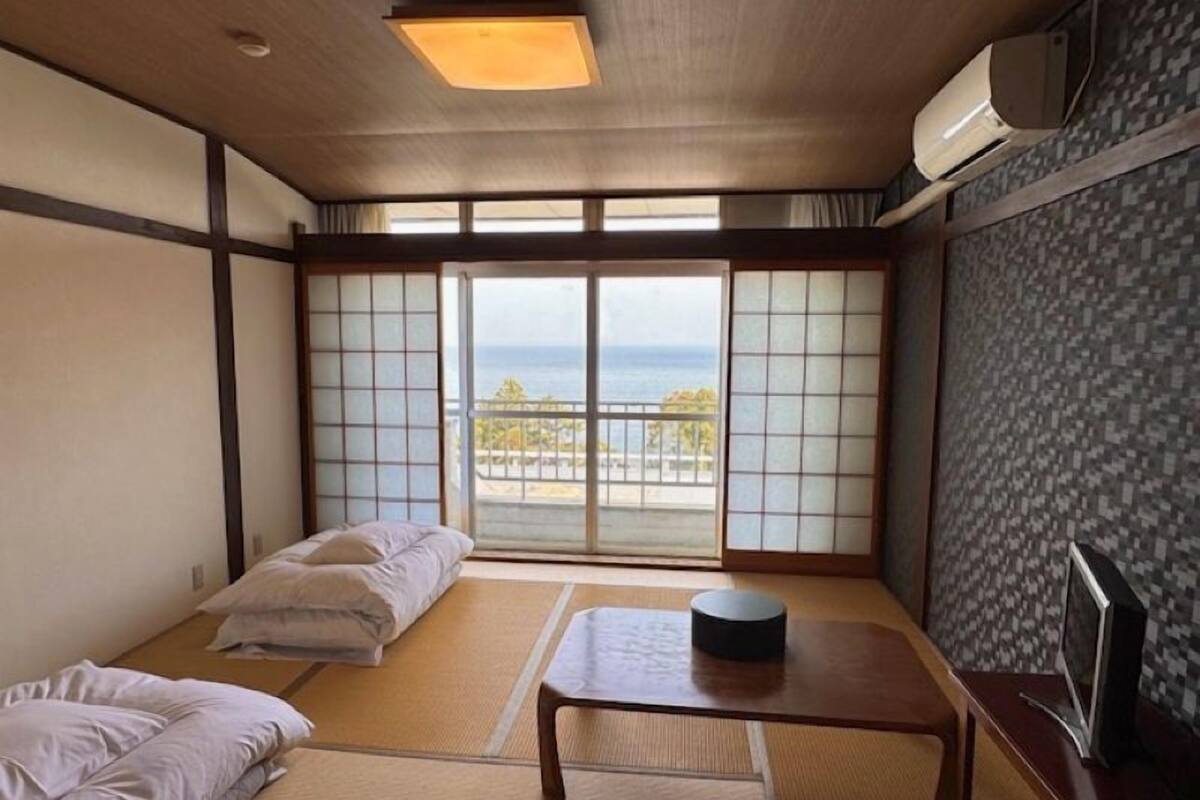 A room with tatami flooring, a futon bed, a small table, a TV, and a view of the sea.