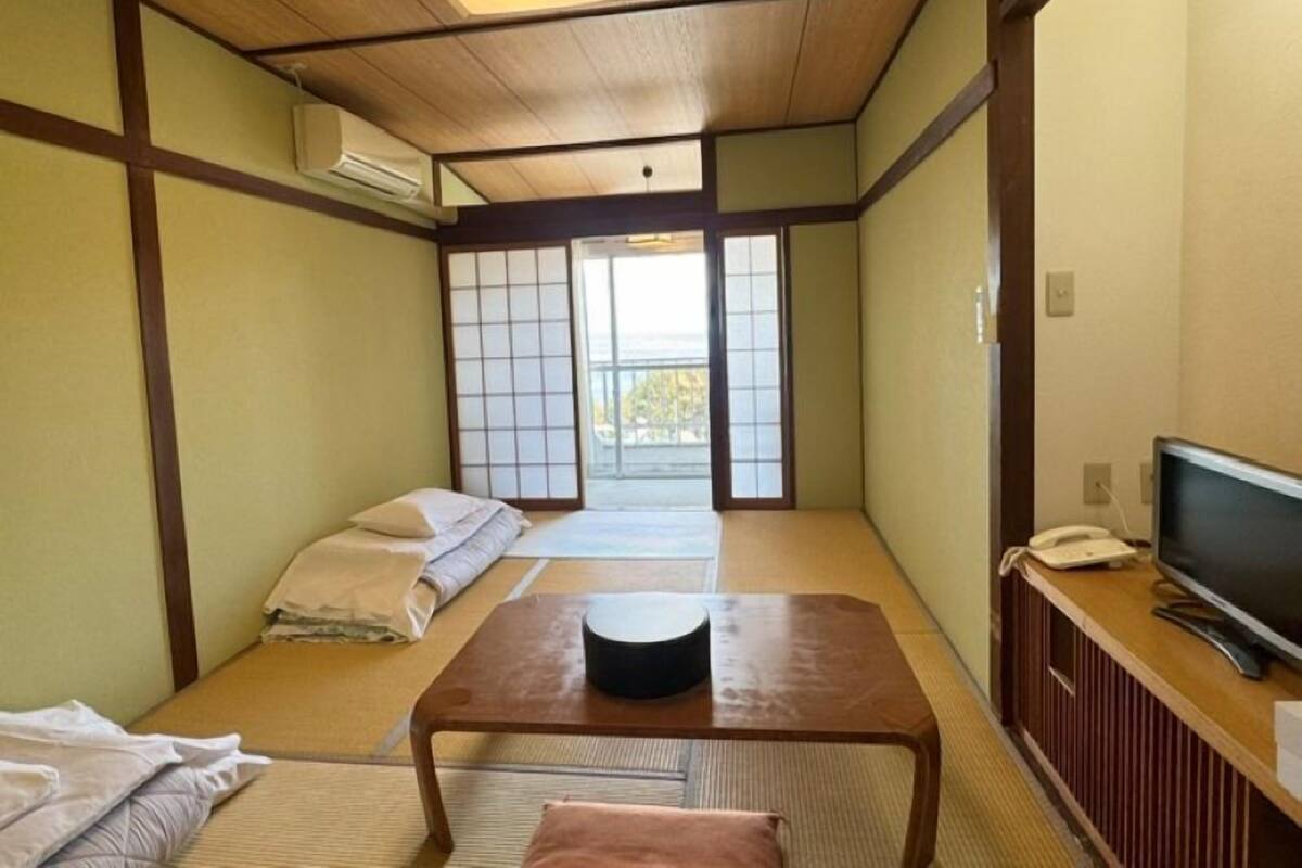 A traditional Japanese room with tatami flooring, a low table, a bed with pillows, a TV, and a sliding door leading to a balcony.