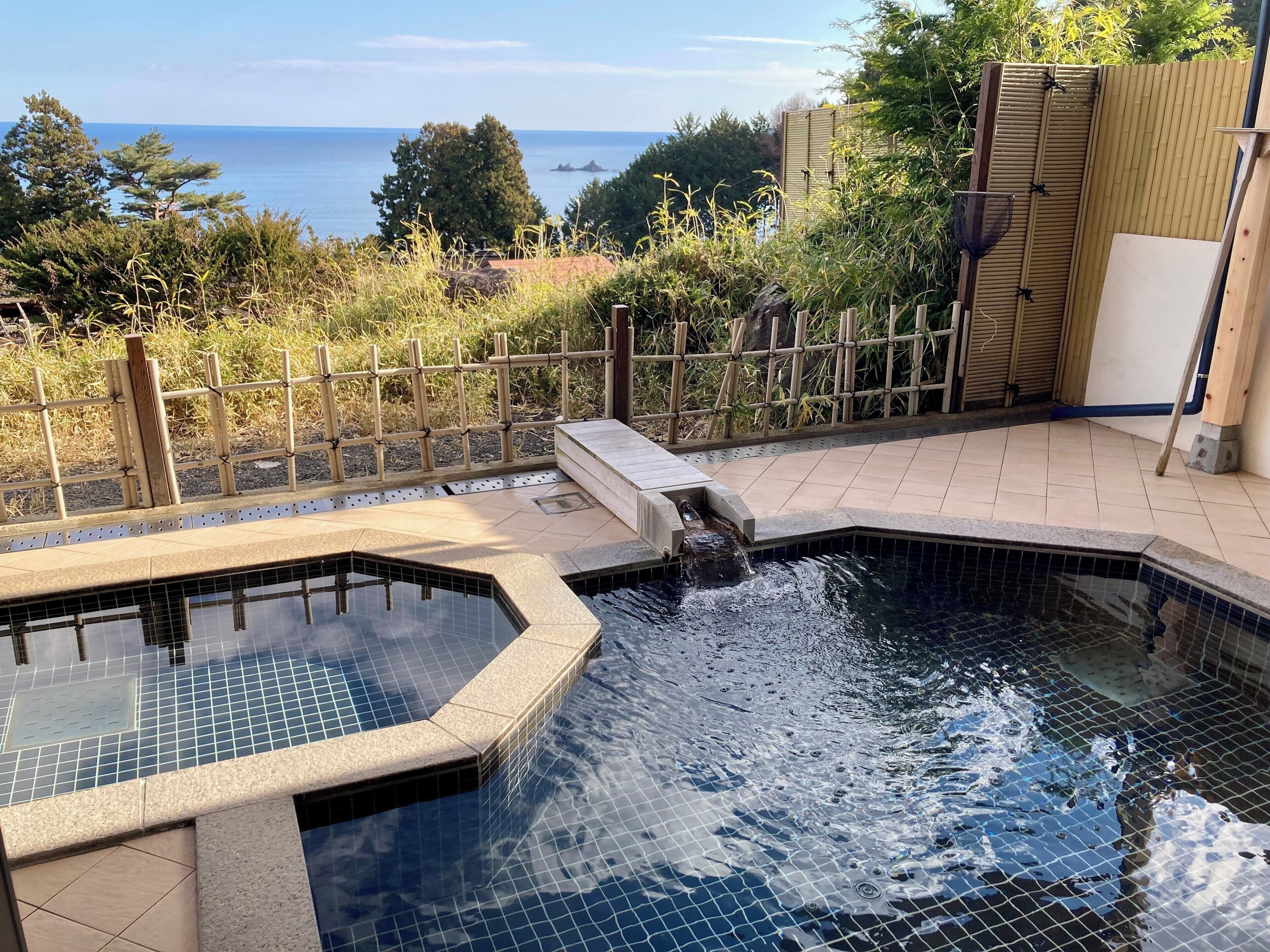 A swimming pool with a waterfall feature, surrounded by a bamboo fence and a view of the sea.
