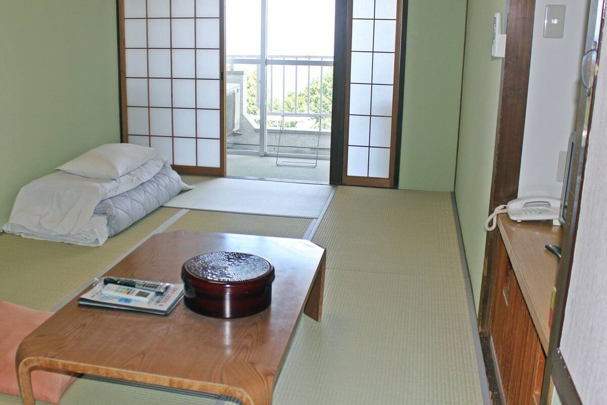 A traditional Japanese room with tatami flooring, a low wooden table with a decorative item, a bed with white bedding, and a sliding door leading to a balcony.