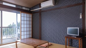 Una habitación con suelo de tatami, una mesa baja, un televisor sobre un mueble y una puerta corrediza que da a un balcón.