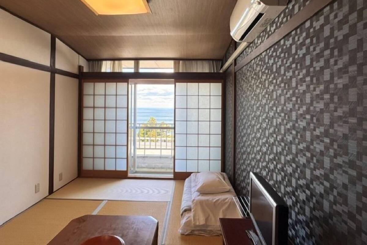 A traditional Japanese room with tatami flooring, a sliding shoji door, a bed with white bedding, and a view of the sea.