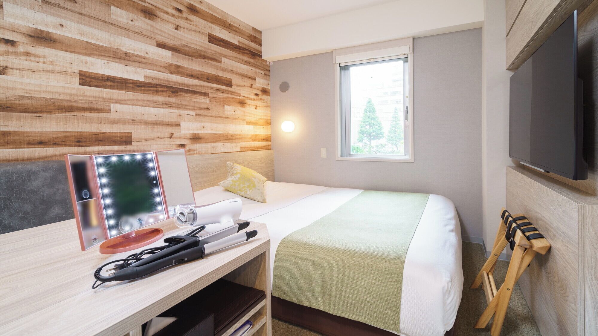 A hotel room with a wooden wall, a bed with a green blanket, a bedside table with a mirror and lights, a TV, and a window with a view of trees.