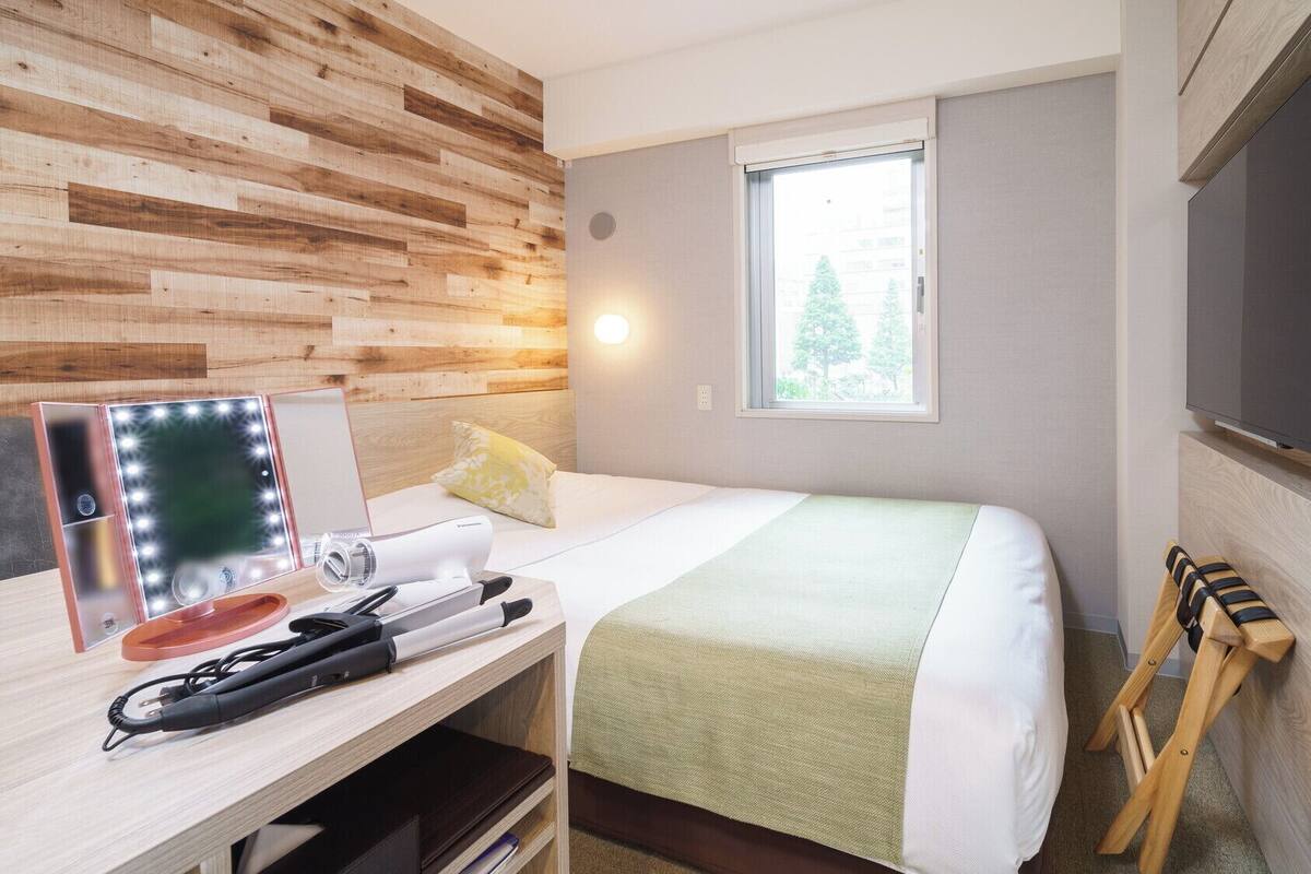Una camera d'albergo con una parete in legno, un letto con coperta verde, un comodino con specchio e luci, una televisione e una finestra vista alberi.