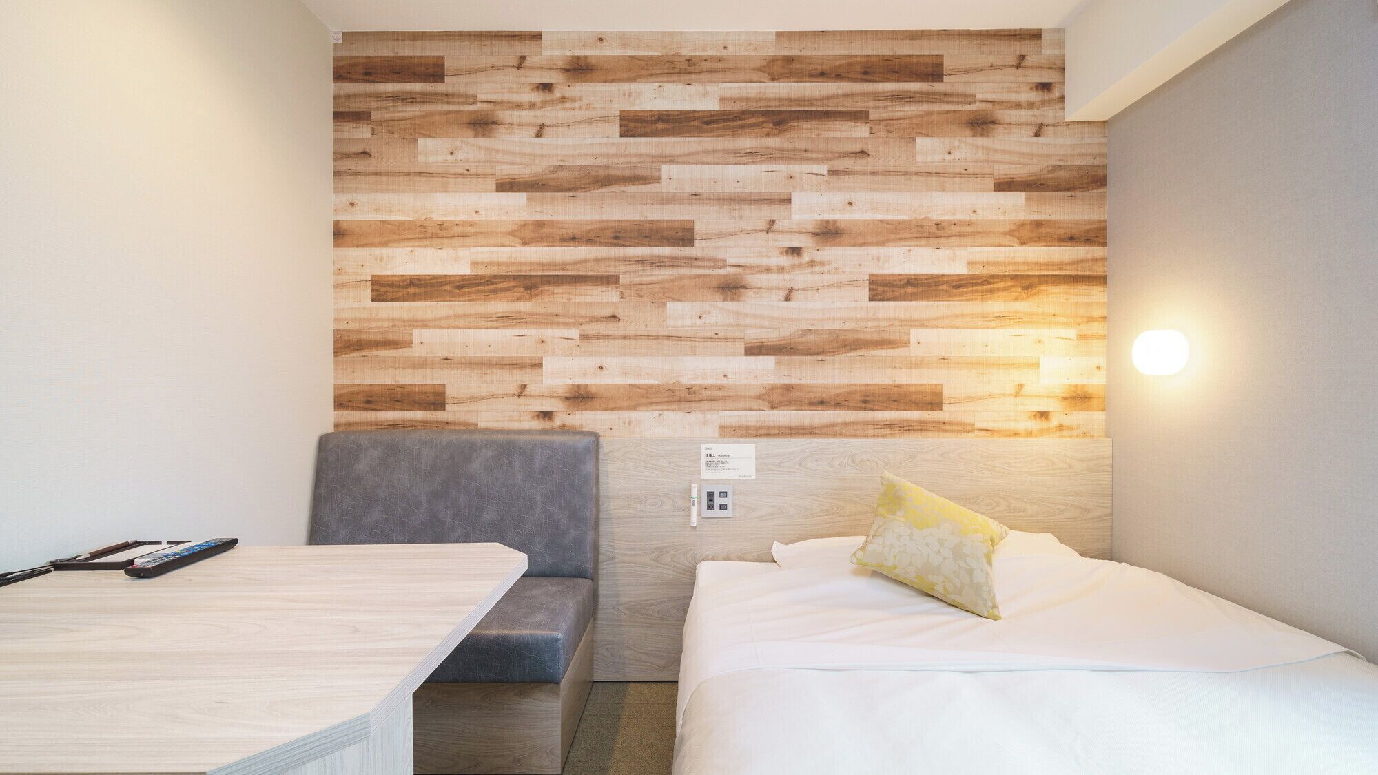 A room with a wooden wall panel, a bed with white bedding, a gray sofa, and a small table with a remote control.