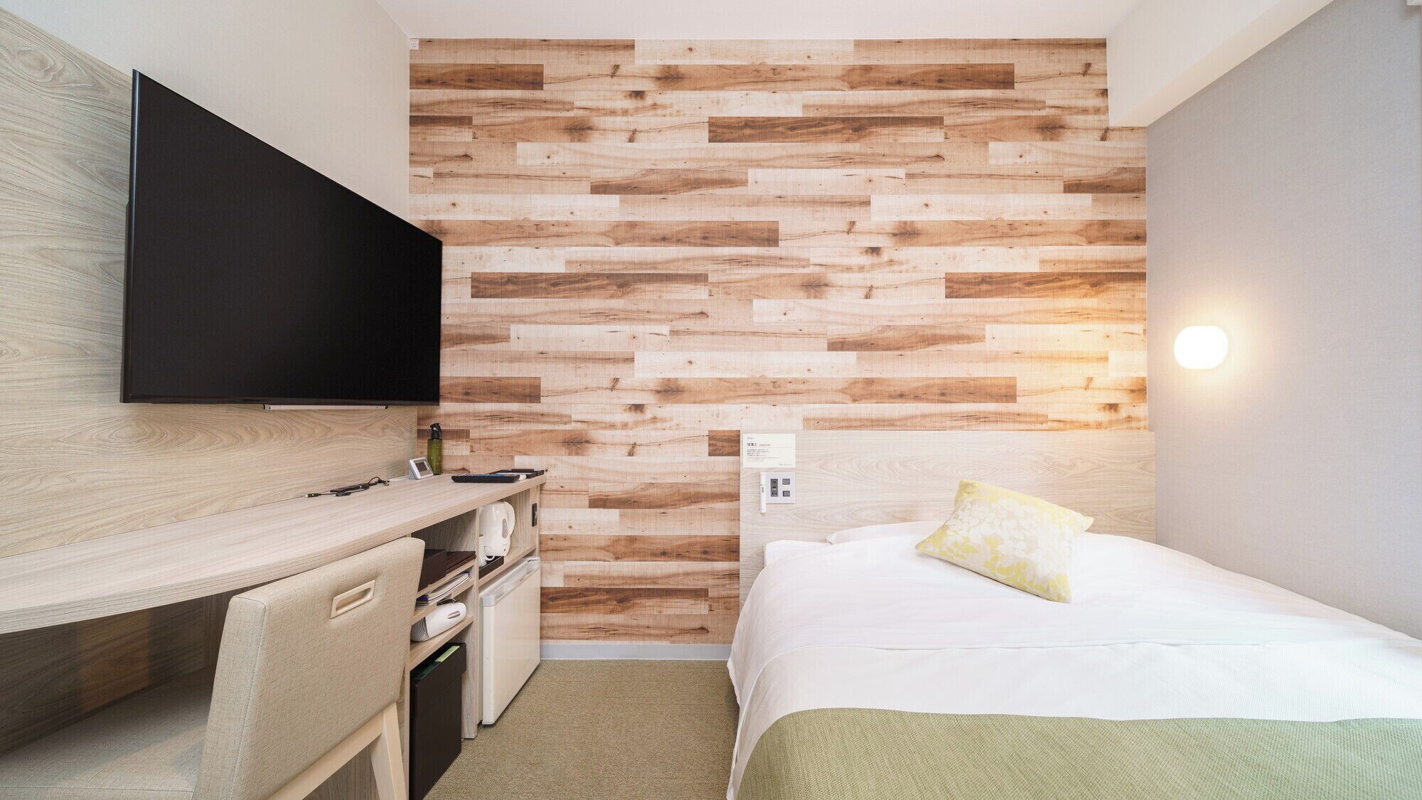 A hotel room with a wooden wall panel, a flat-screen TV, a desk with a chair, a bed with a pillow, and a bedside table.