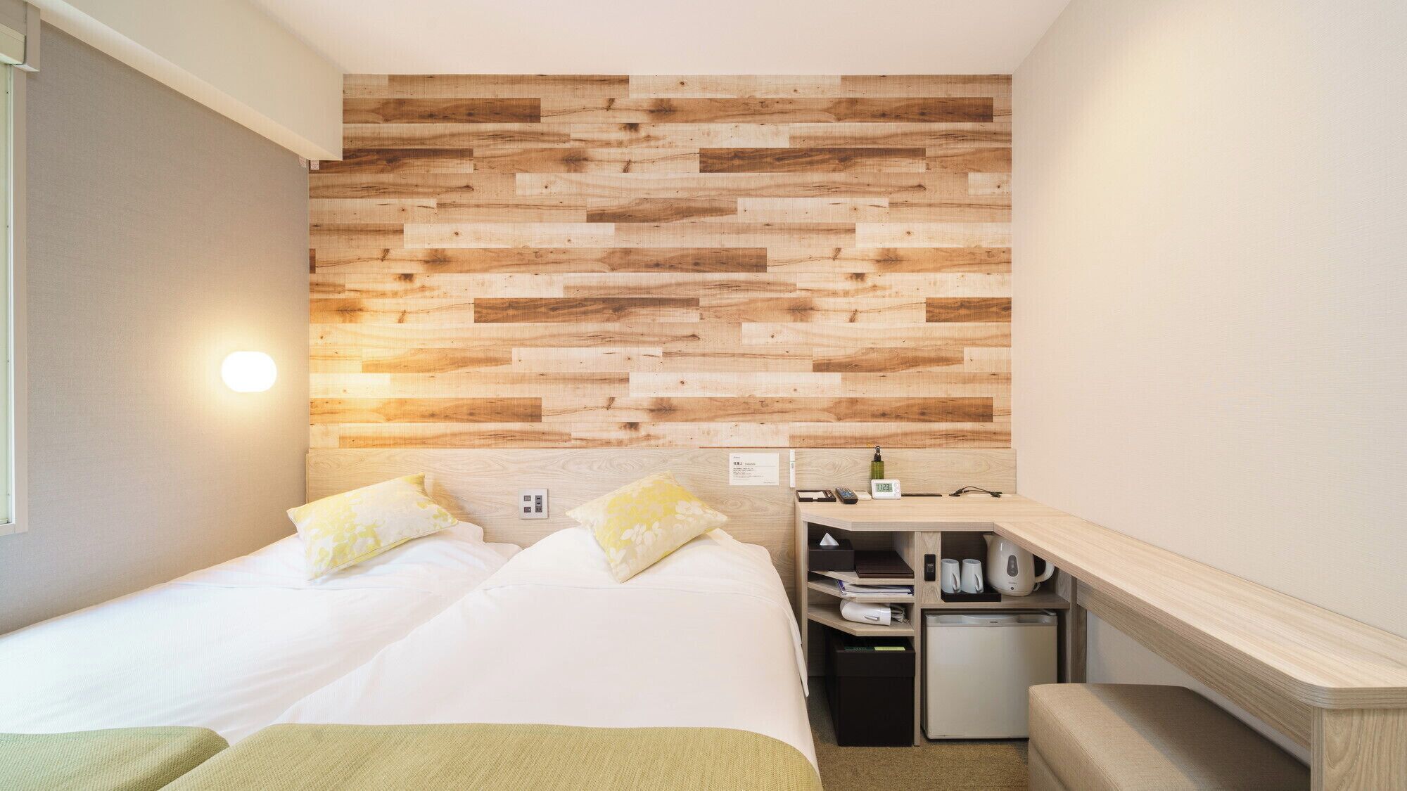 A bedroom with a wooden wall panel, a bed with pillows, a bedside table with a clock, and a small kitchenette.