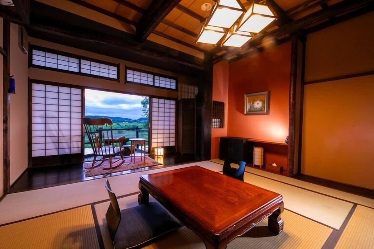 Una stanza tradizionale giapponese con pavimento in tatami, un tavolino, una poltrona a dondolo e vista su un paesaggio attraverso uno schermo scorrevole.