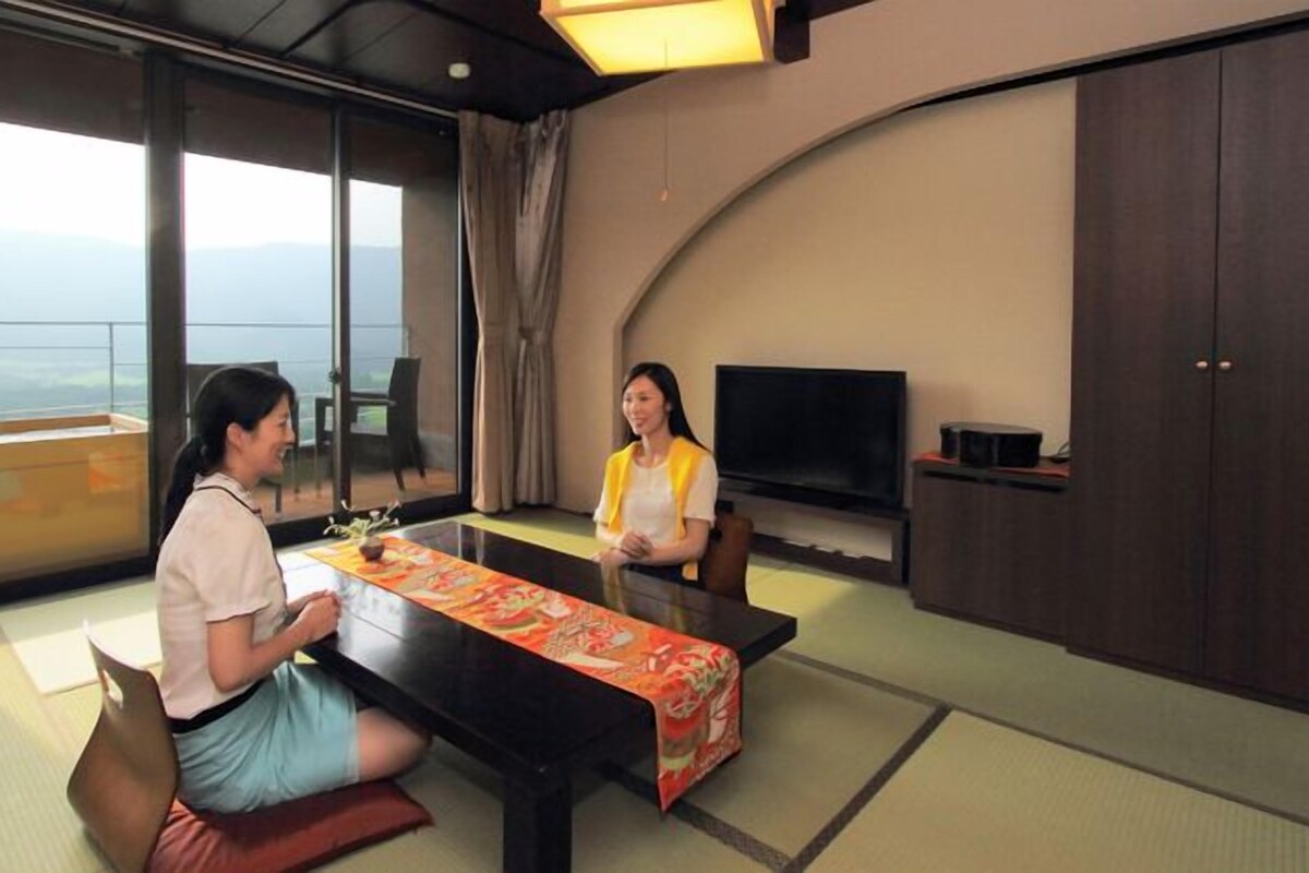 A room with tatami flooring, a low table with a colorful tablecloth, a television, and a view of a mountainous landscape through large windows.
