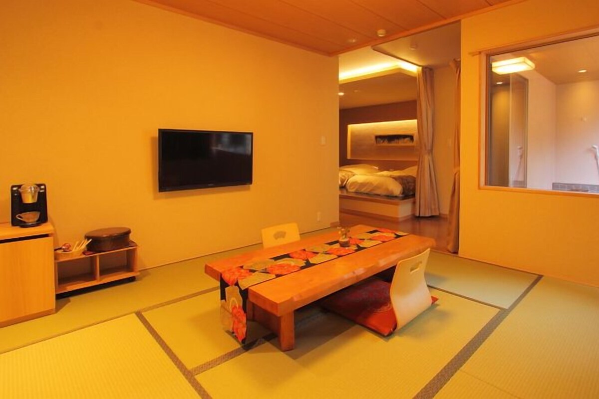 A traditional Japanese-style room with tatami flooring, a low wooden table, a television, and a sliding door leading to a bedroom.